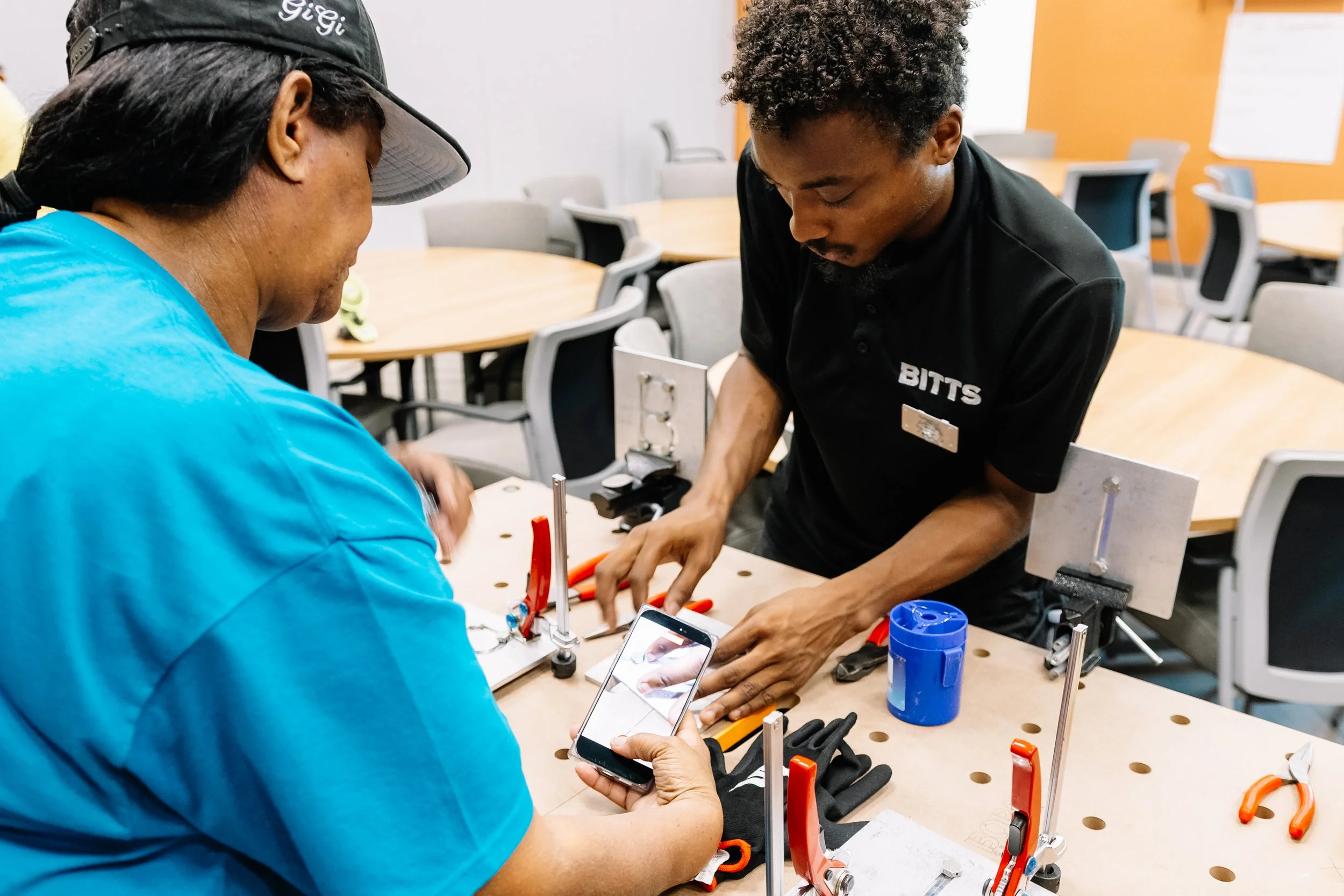 A BITTS instructor guides an attendee through a hands-on building activity using tools and materials at a workstation during Chakira’s Cookout.