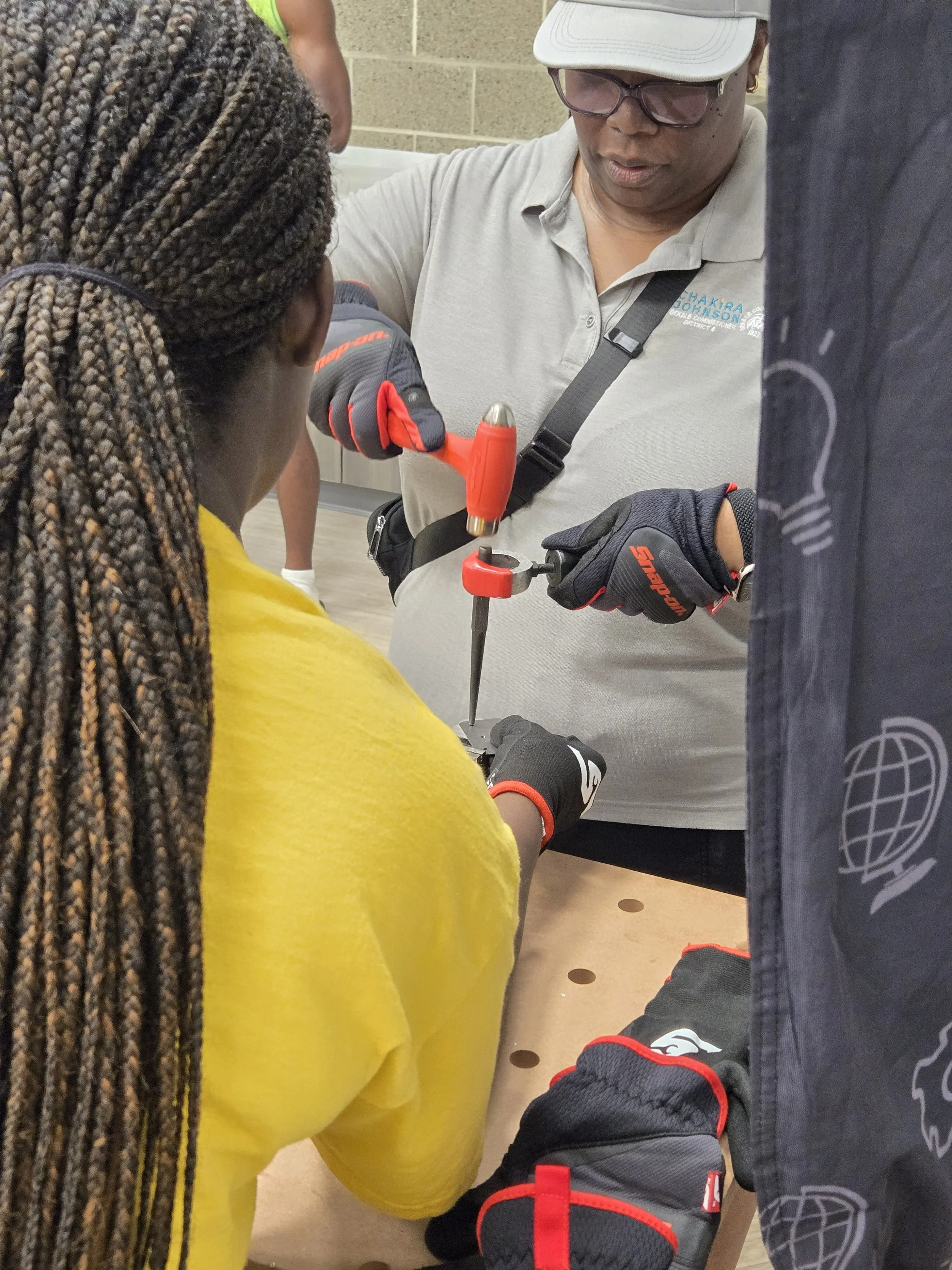 Commissioner Chakira Johnson, wearing work gloves, strikes a metal punch with a red mallet while a volunteer steadies the project.