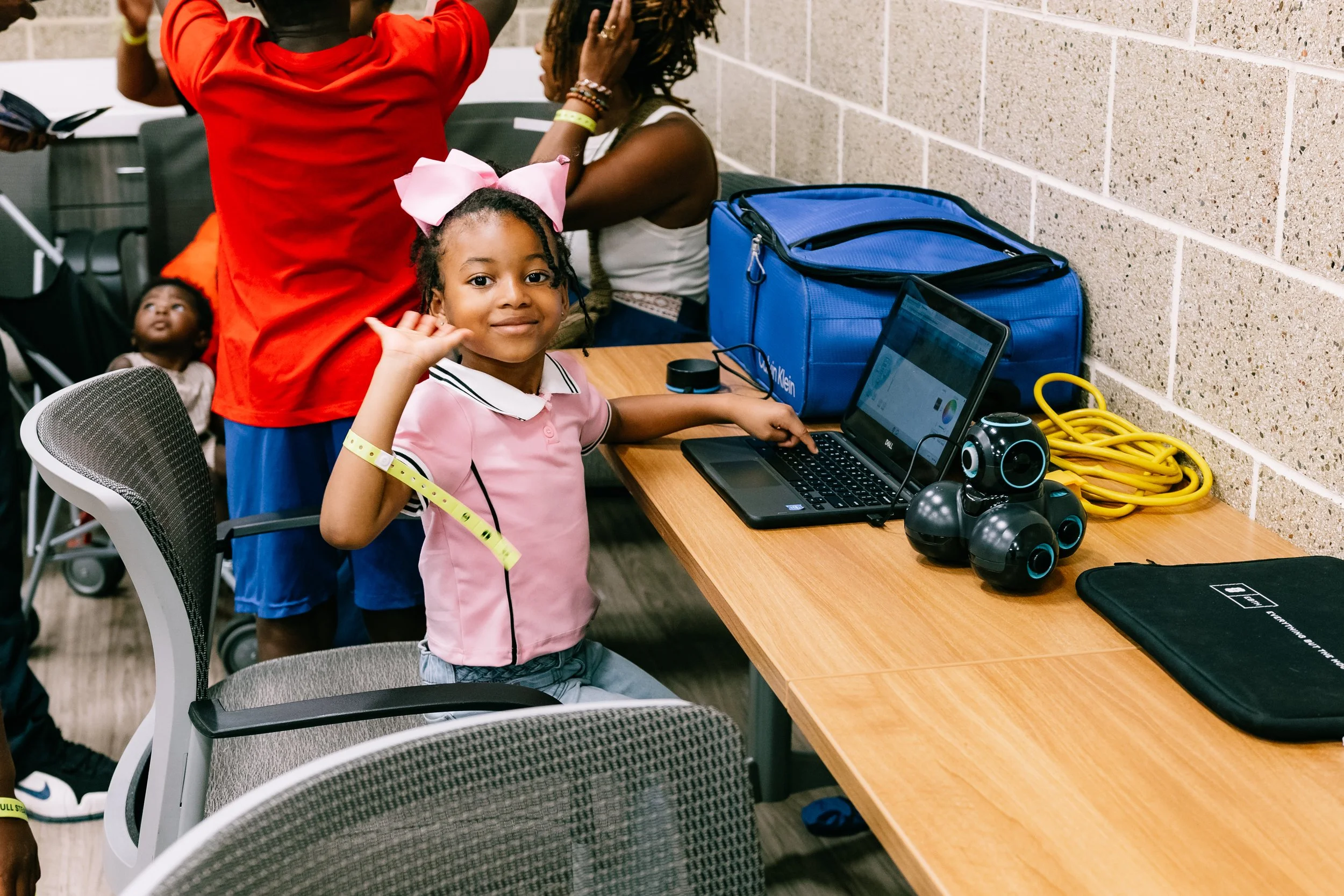 Young attendee smiles and waves at the camera while seated beside a laptop and two small robots during a hands-on robotics activity at Chakira’s Cookout.