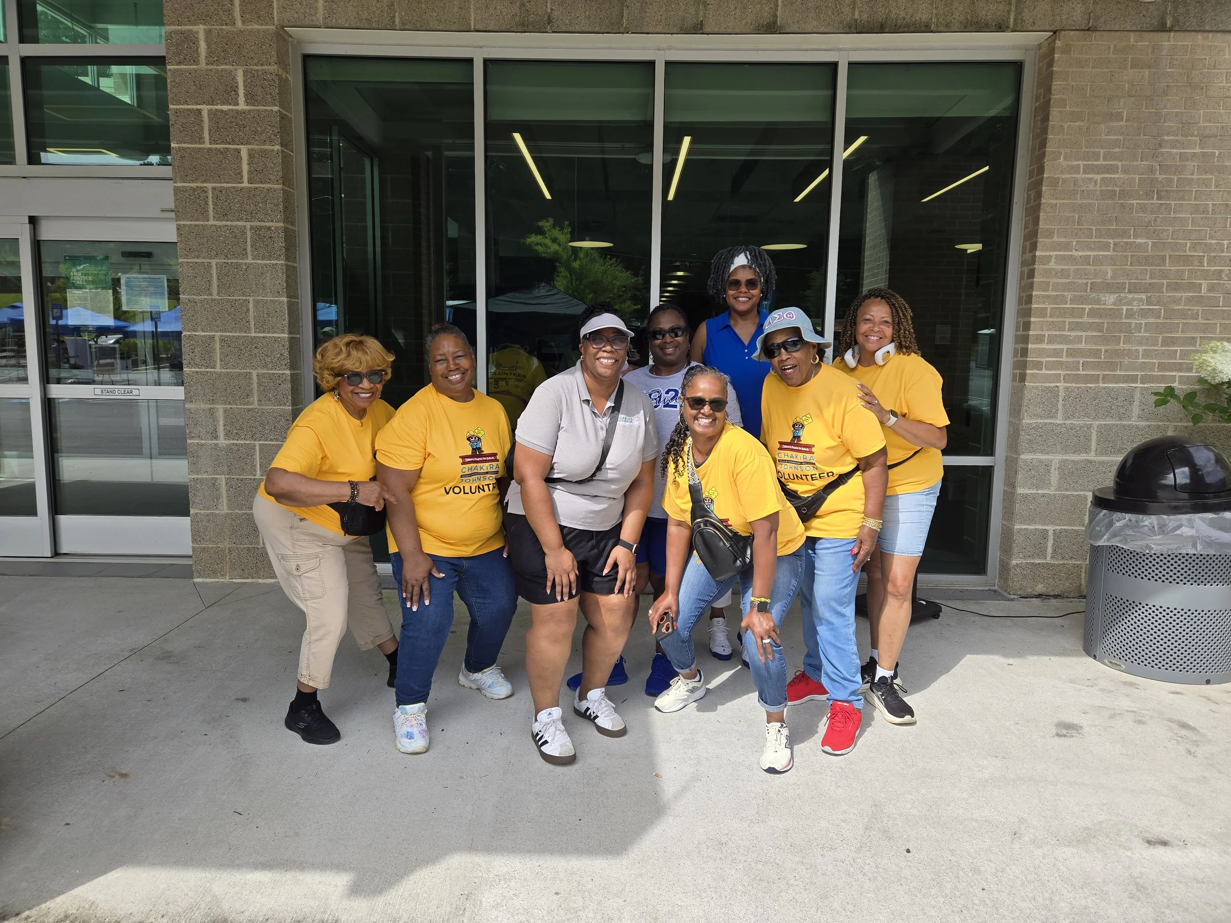 Commissioner Chakira Johnson, wearing a light gray polo, poses outdoors with seven Chakira’s Cookout volunteers, wearing branded yellow Chakira's Cookout Volunteer T-shirts.