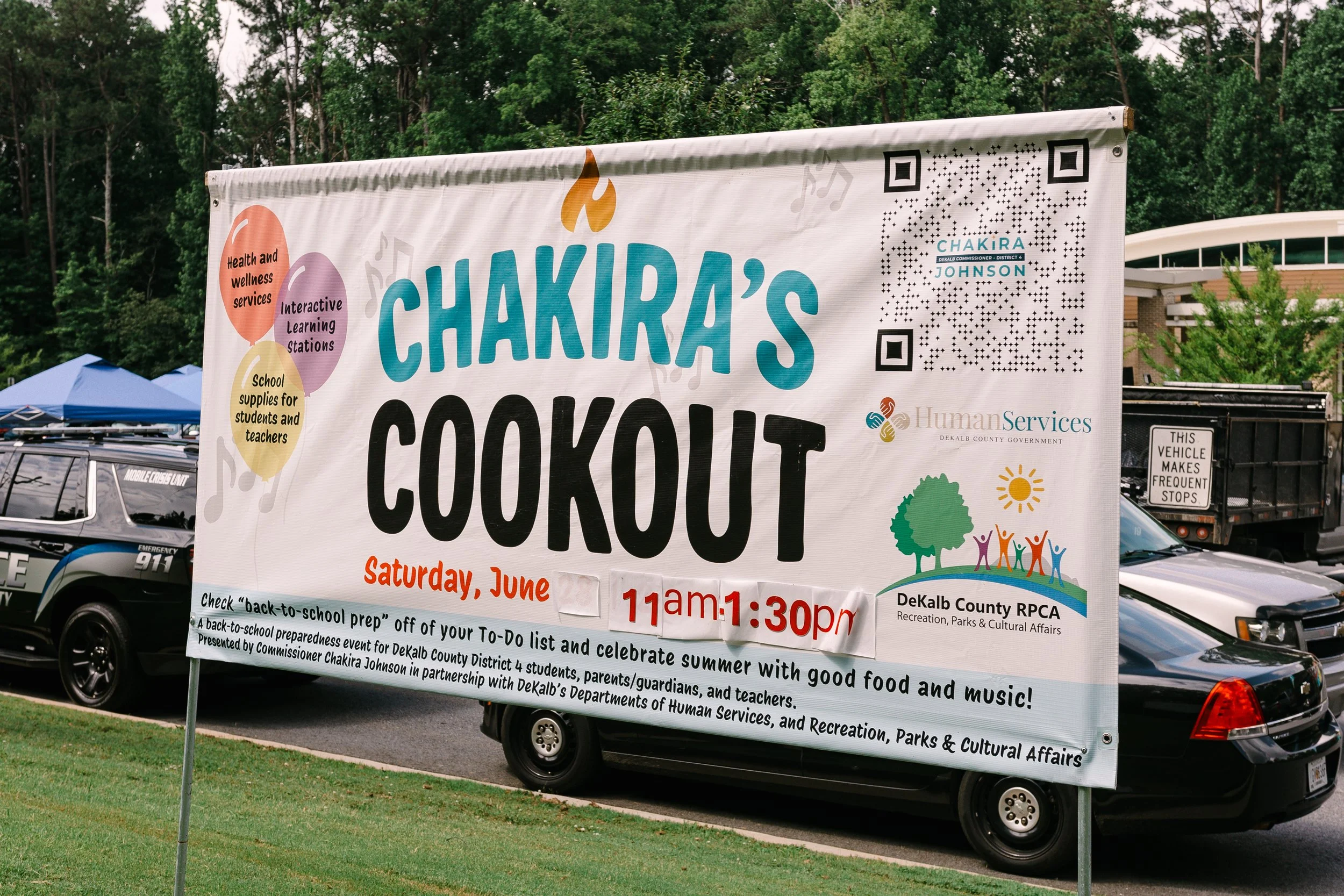 Chakira’s Cookout event banner displayed outdoors, featuring the event name and information about back-to-school resources, interactive learning stations, health and wellness services, food, parks and music. DeKalb County public safety vehicles are p