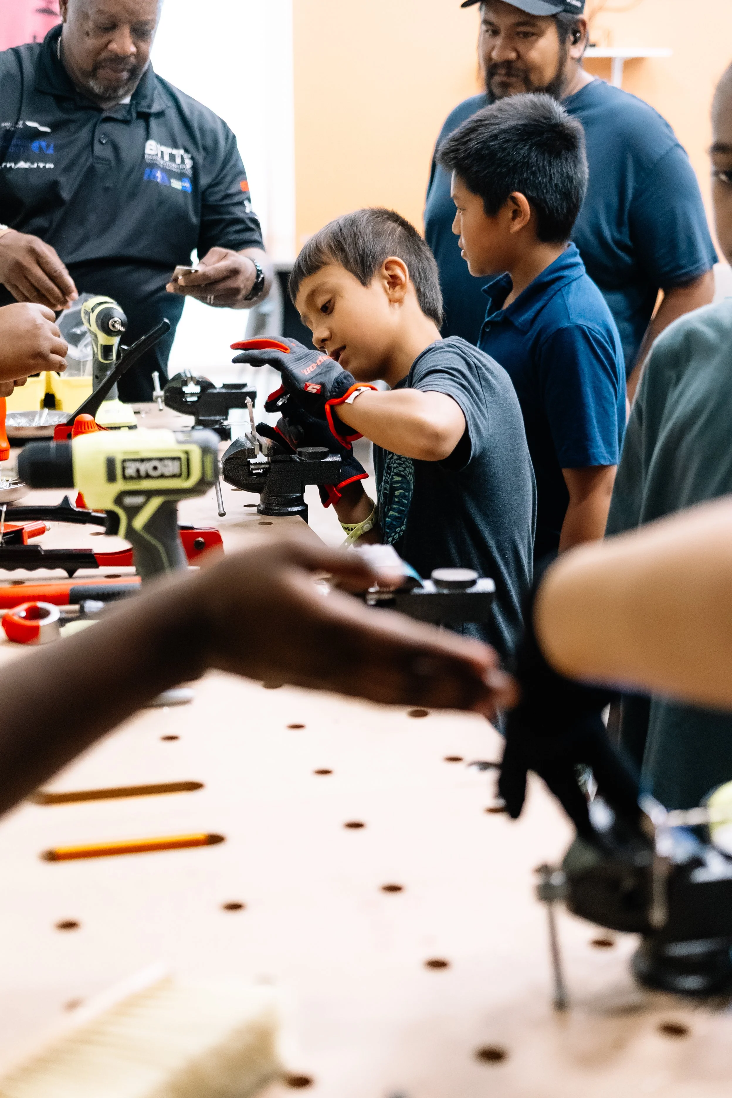 Young attendee concentrates on a hands-on building project using a bench vise while a BITTS instructor and other participants look on during Chakira’s Cookout.