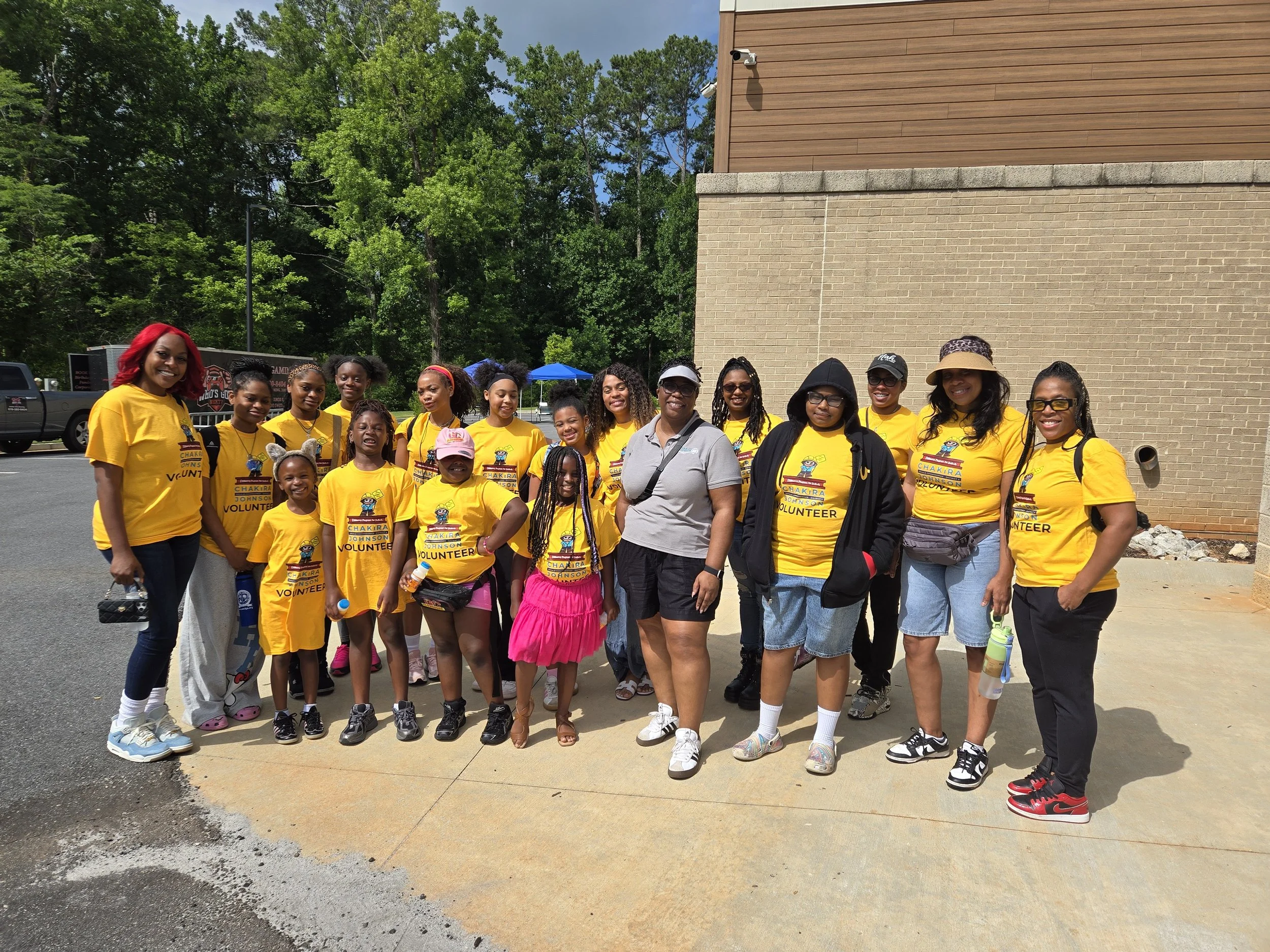 Commissioner Chakira Johnson, wearing a light gray polo, poses outdoors with a large group of volunteers, most wearing branded yellow Chakira’s Cookout Volunteer t-shirts.