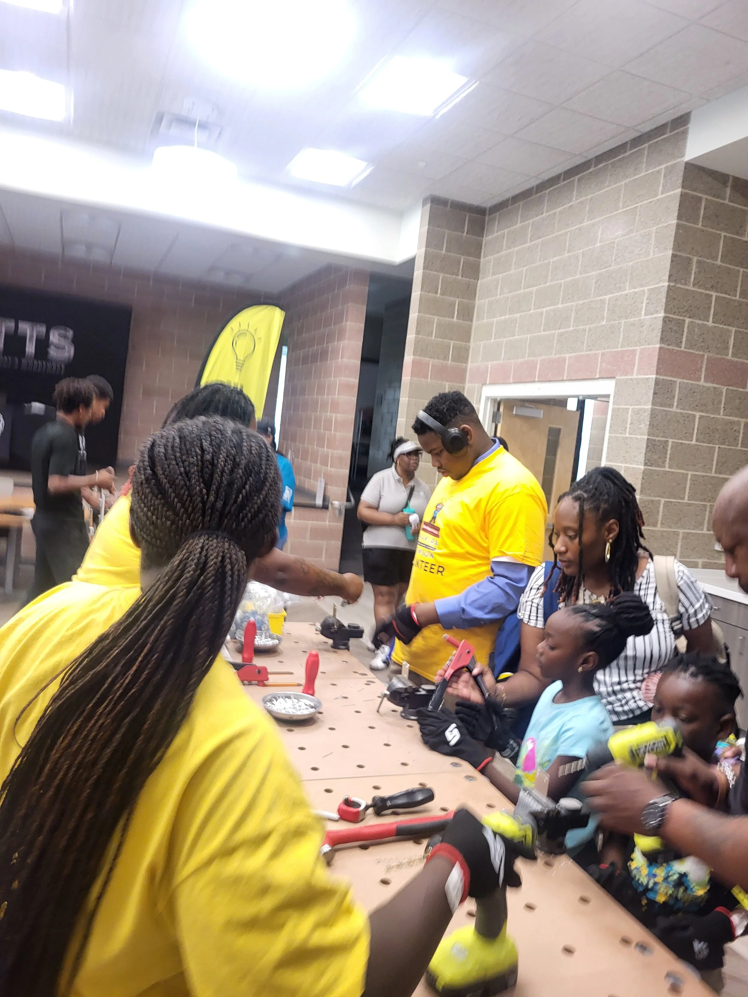 Two children explore drills and hand tools at a Flying Classroom workbench with guidance from family members and volunteers.