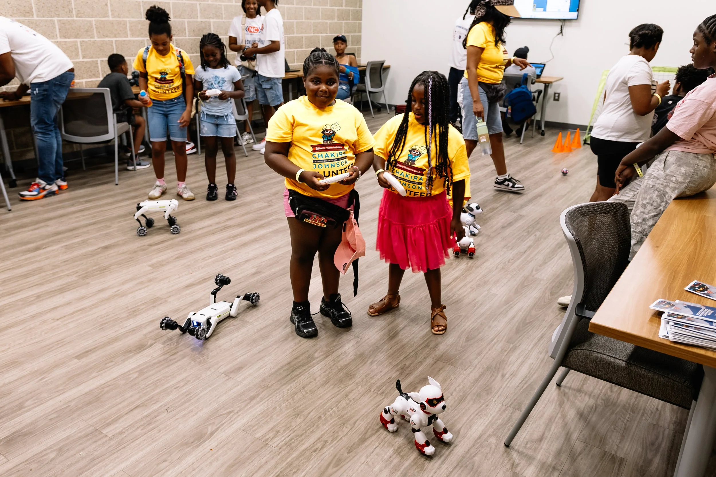 Two children operate small robotic dogs during an interactive robotics activity at Chakira’s Cookout, while other children and volunteers participate nearby.