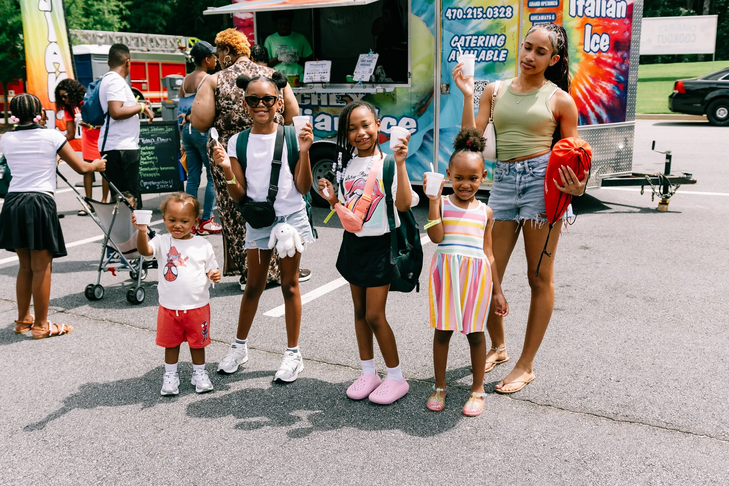 Four children smile and hold up cups of Italian ice while standing near Michey’s Italian Ice food trailer during Chakira’s Cookout.
