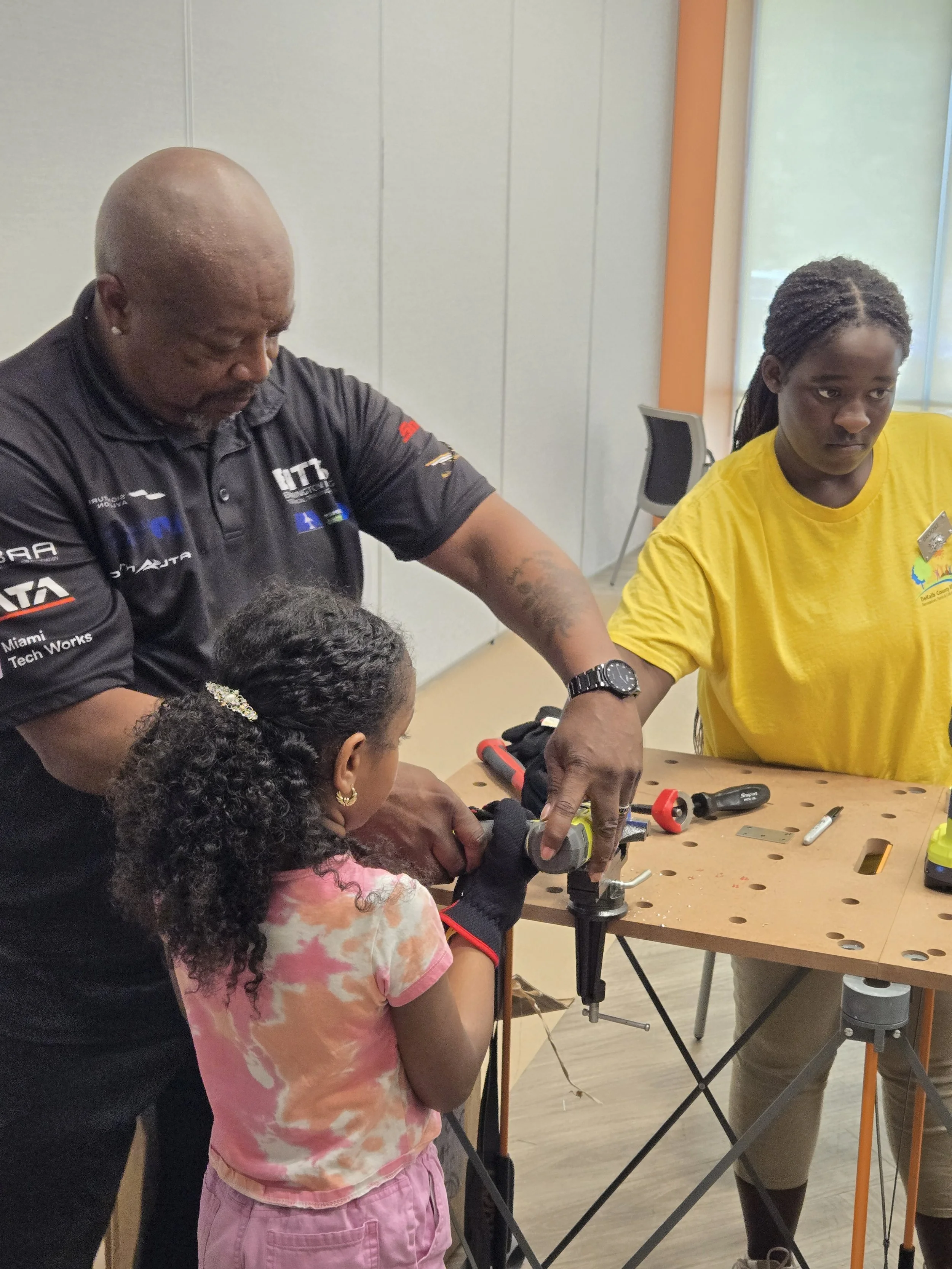 A Flying Classroom instructor guides a young girl wearing work gloves as she uses a power drill on a small metal piece secured in a table vise.