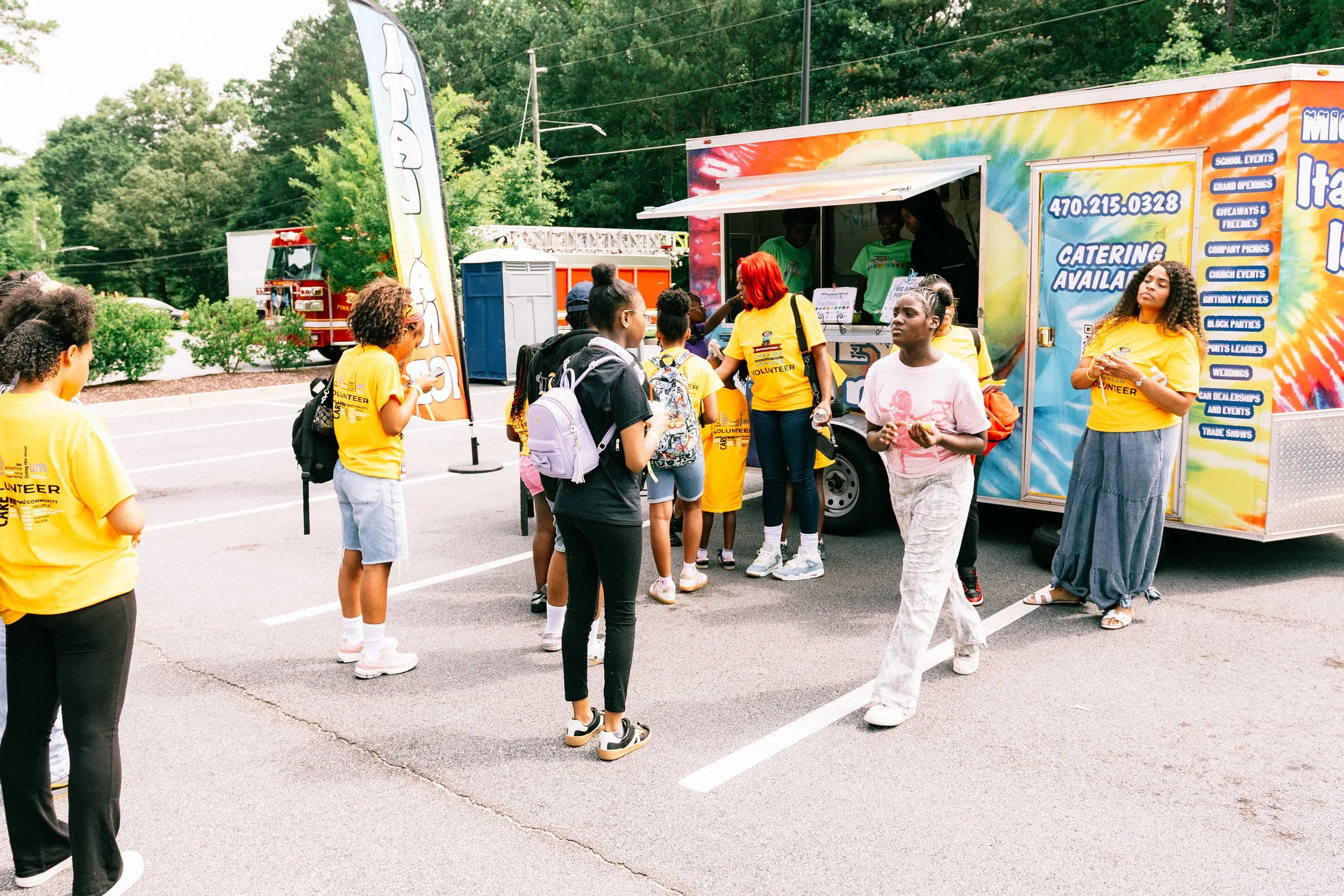Chakira’s Cookout attendees and volunteers gather near Michey’s Italian Ice food trailer, with several volunteers wearing bright yellow event shirts as guests wait to be served.