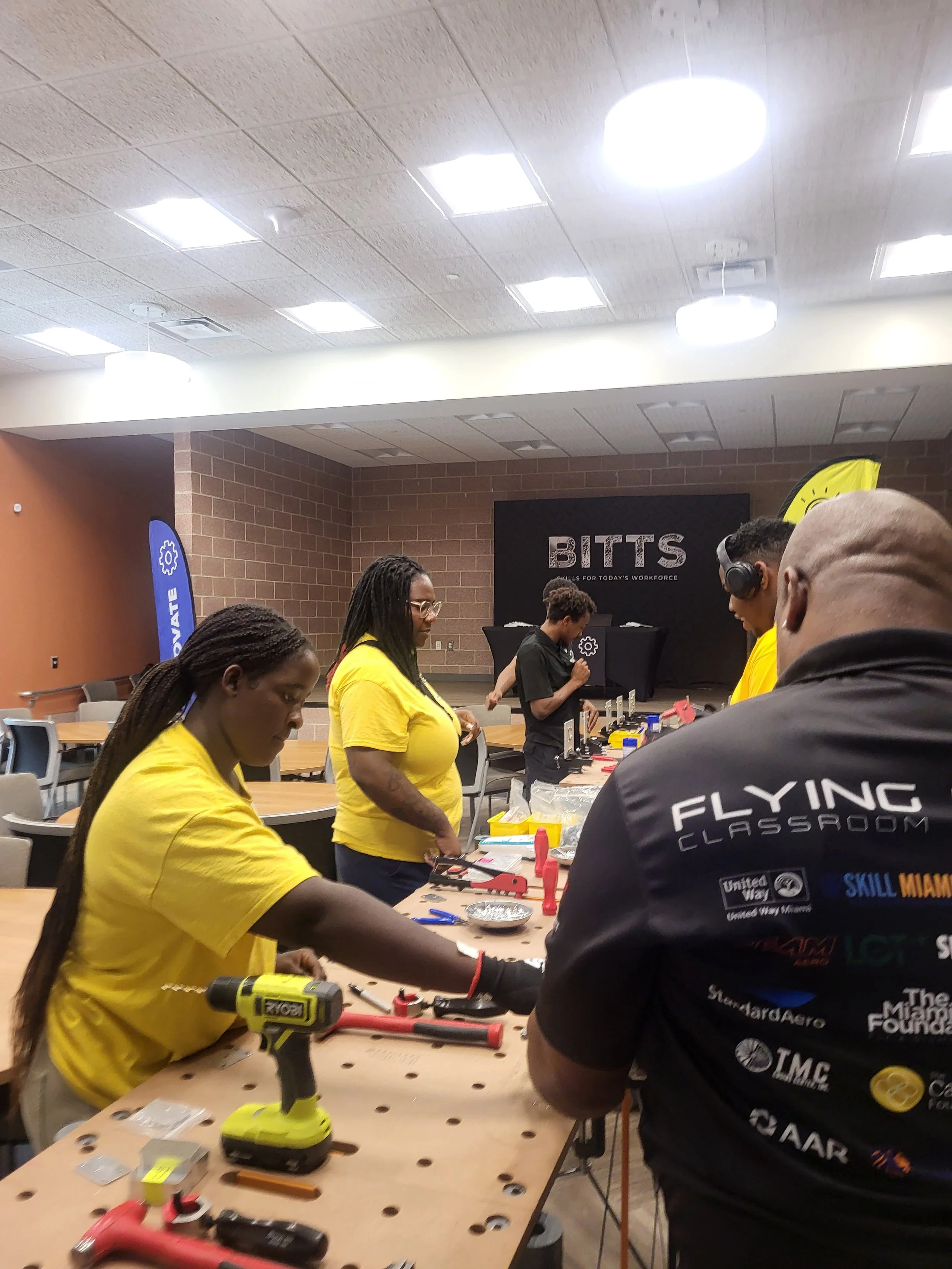 Chakira’s Cookout volunteers and Flying Classroom instructors assist a participant wearing protective eyewear and hearing protection at a tool-filled workbench.