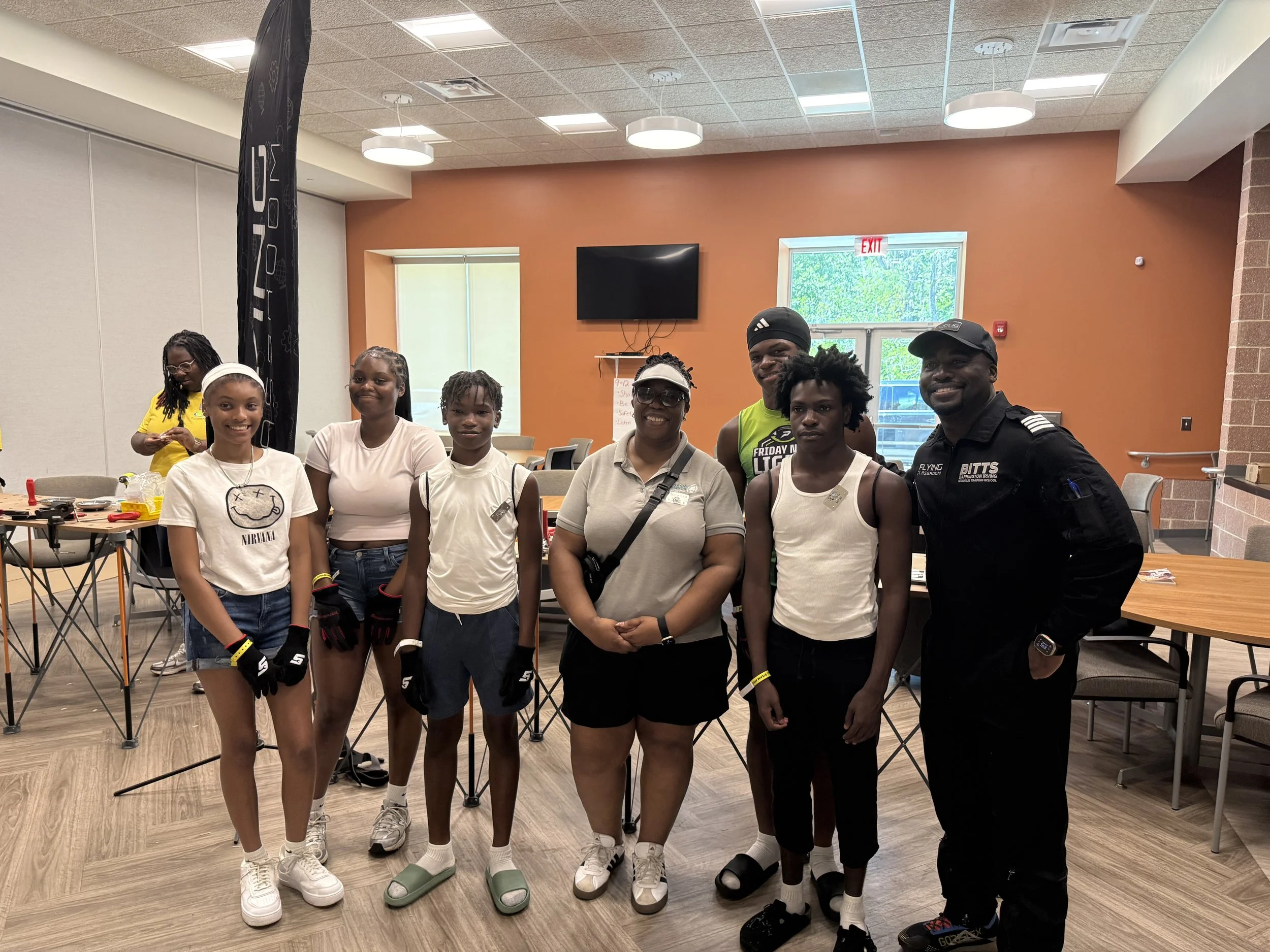Commissioner Chakira Johnson and Captain Barrington Irving pose with five young participants inside the Flying Classroom activity room, with workstations visible behind them.