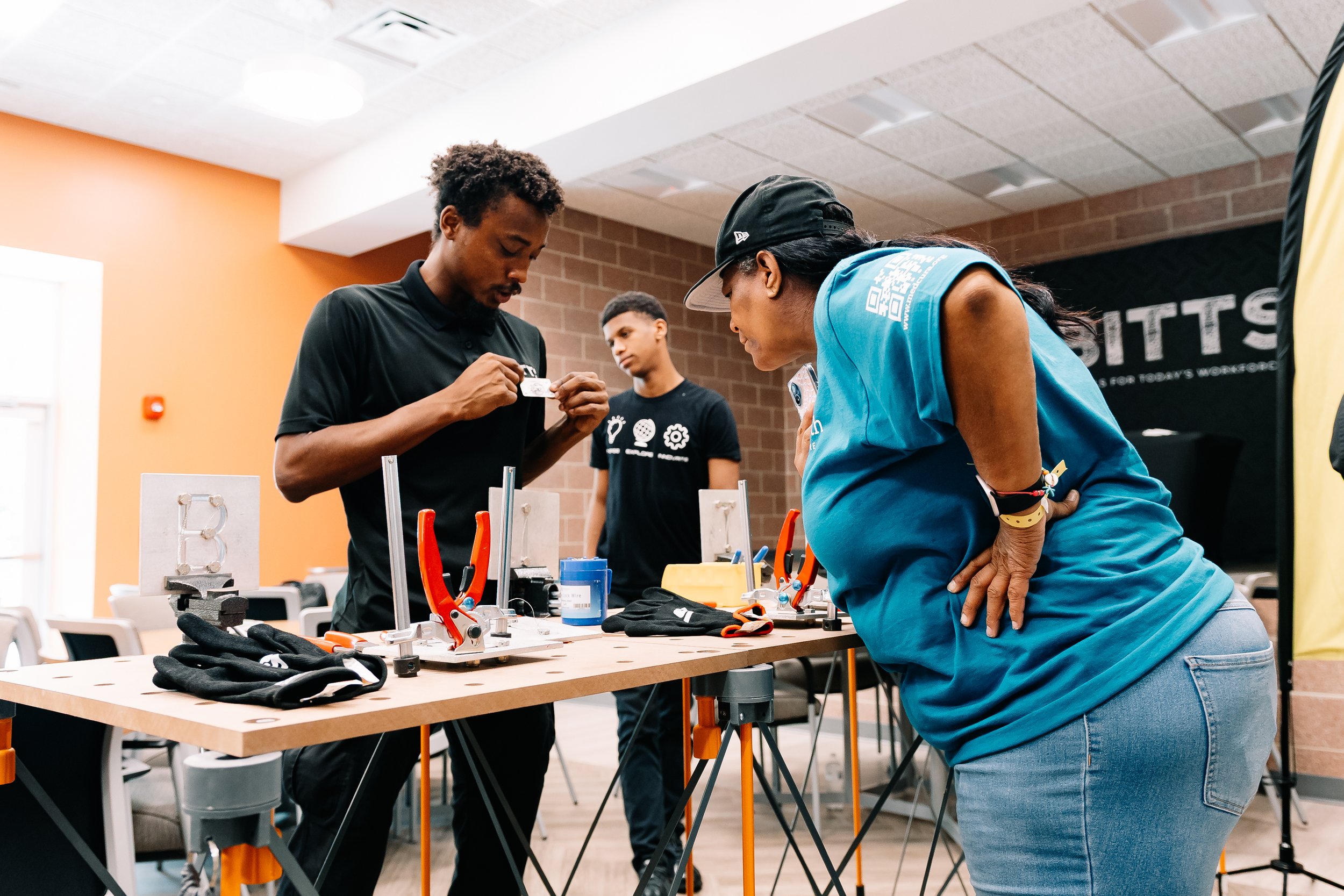 A BITTS instructor demonstrates a hands-on building activity as an attendee watches closely at a tool-equipped workstation during Chakira’s Cookout.