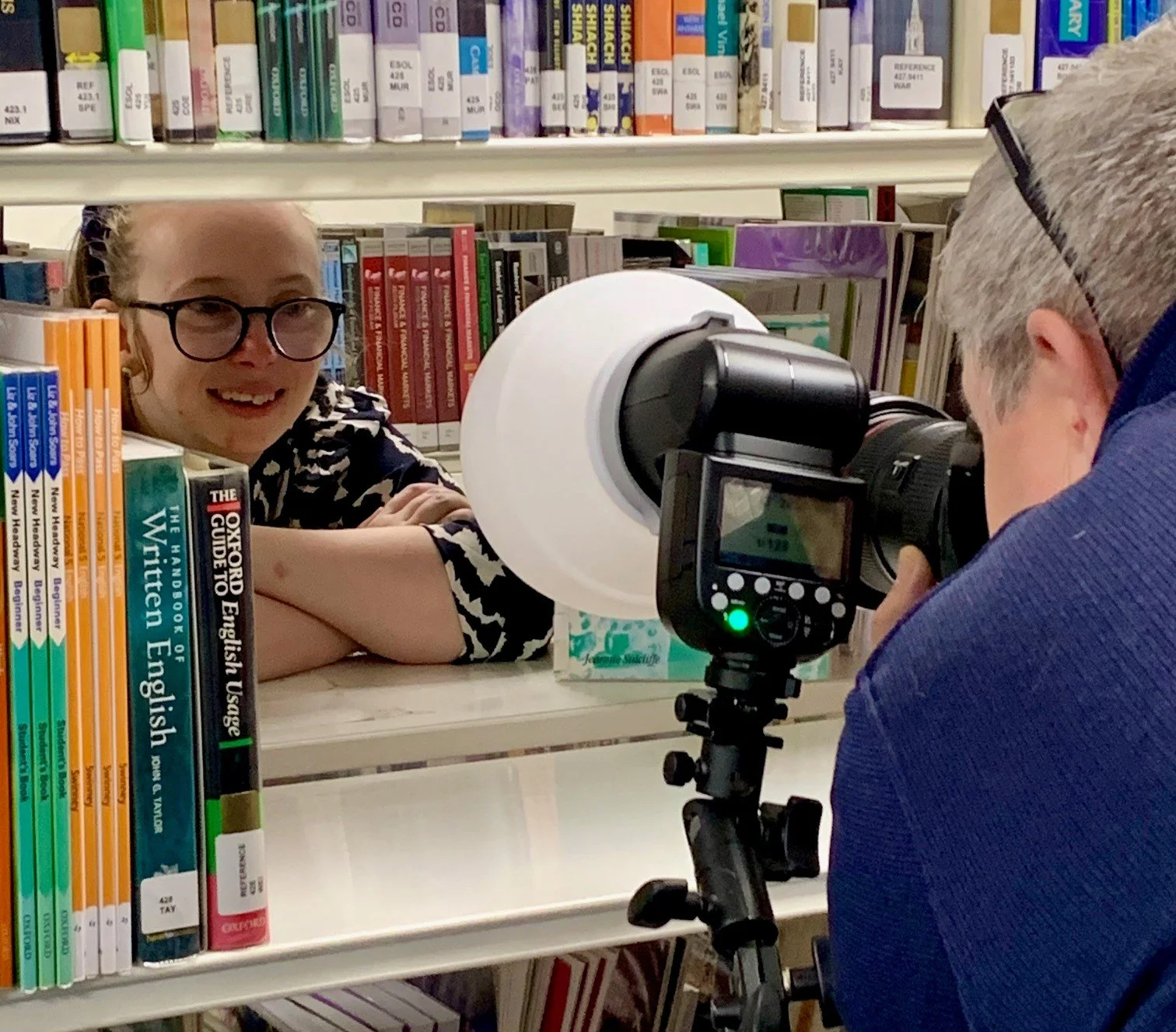 Person being photographed in a library with books on shelves, using a camera with a flash diffuser.