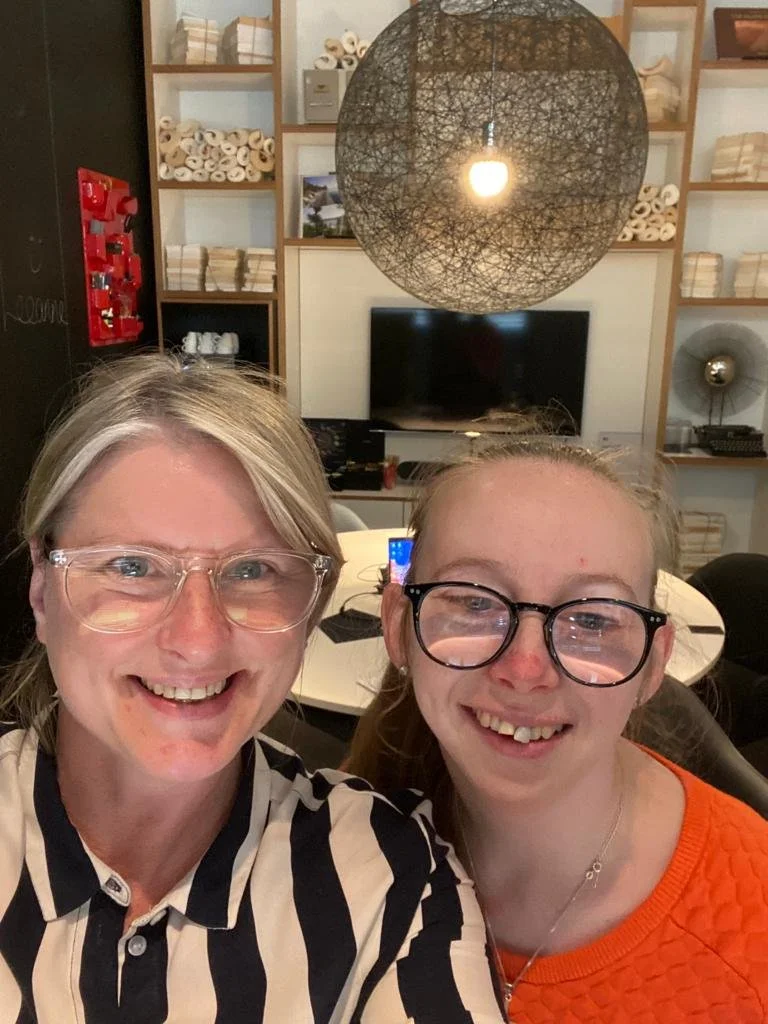 Two people smiling, wearing glasses, in an indoor setting with shelves, a hanging light fixture, and a television in the background.