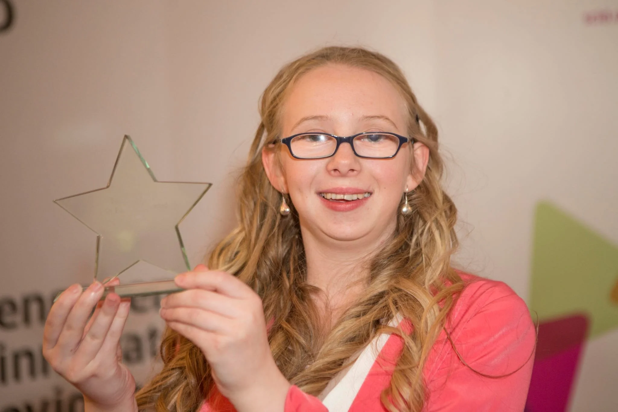 Person holding a star-shaped glass award and smiling, wearing glasses and a pink outfit.