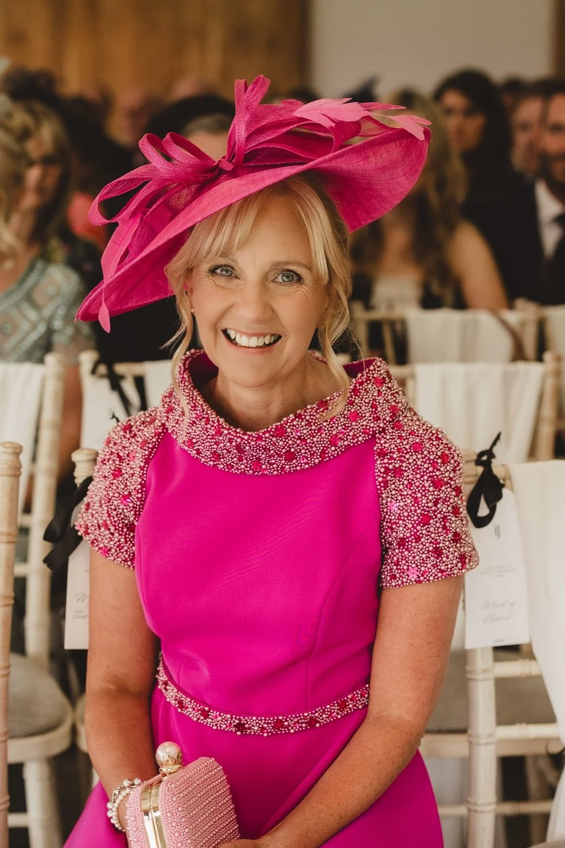 A woman dressed in a vibrant pink dress with intricate beadwork on the shoulders, wearing a large matching pink hat with decorative feathers, smiling at a formal event or gathering.