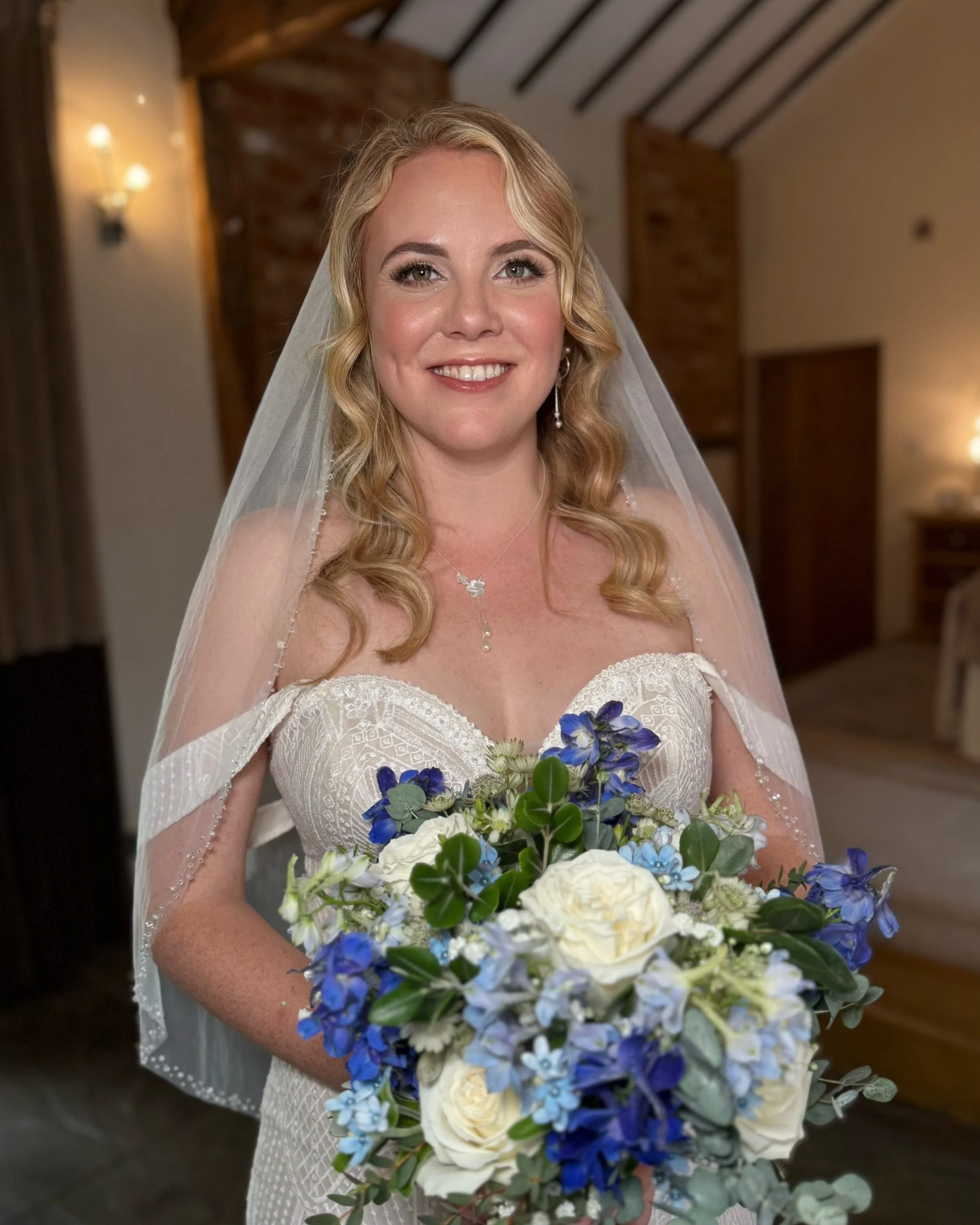 A bride with blonde, wavy hair, wearing a white lace wedding dress and veil, holding a bouquet of white roses, blue flowers, and green foliage, standing indoors with wooden and brick decor.
