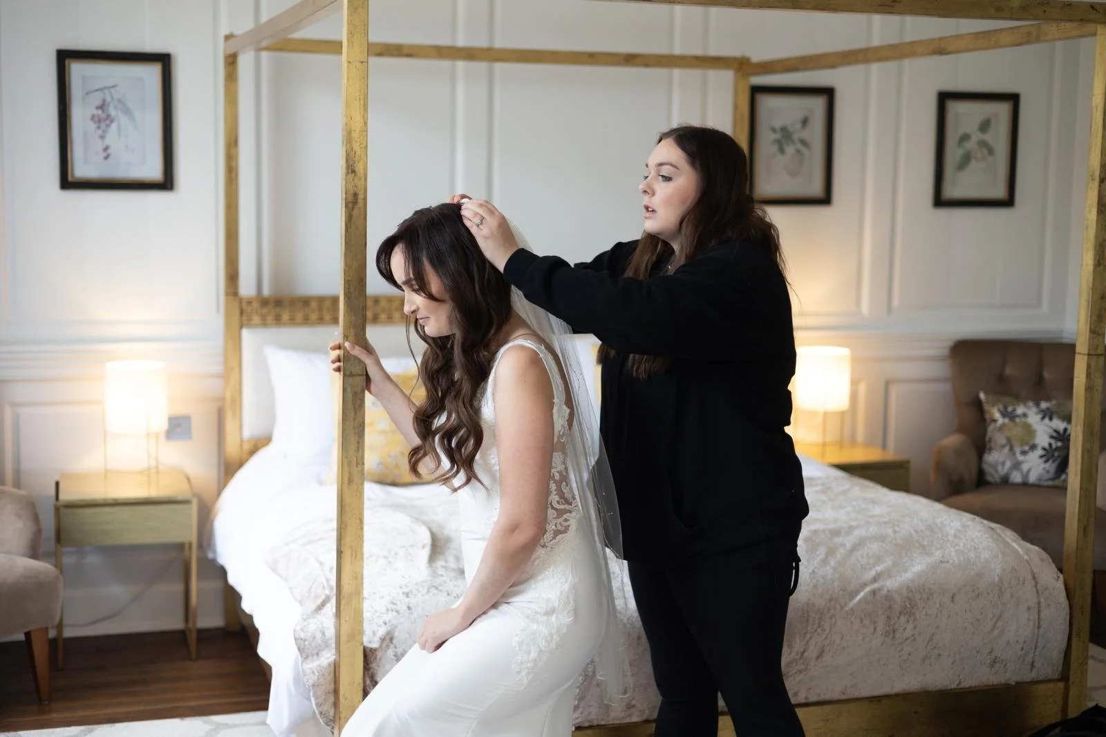 A woman in a wedding dress sits on a bed while another woman helps her with her veil in a bedroom decorated with framed pictures and lamps.