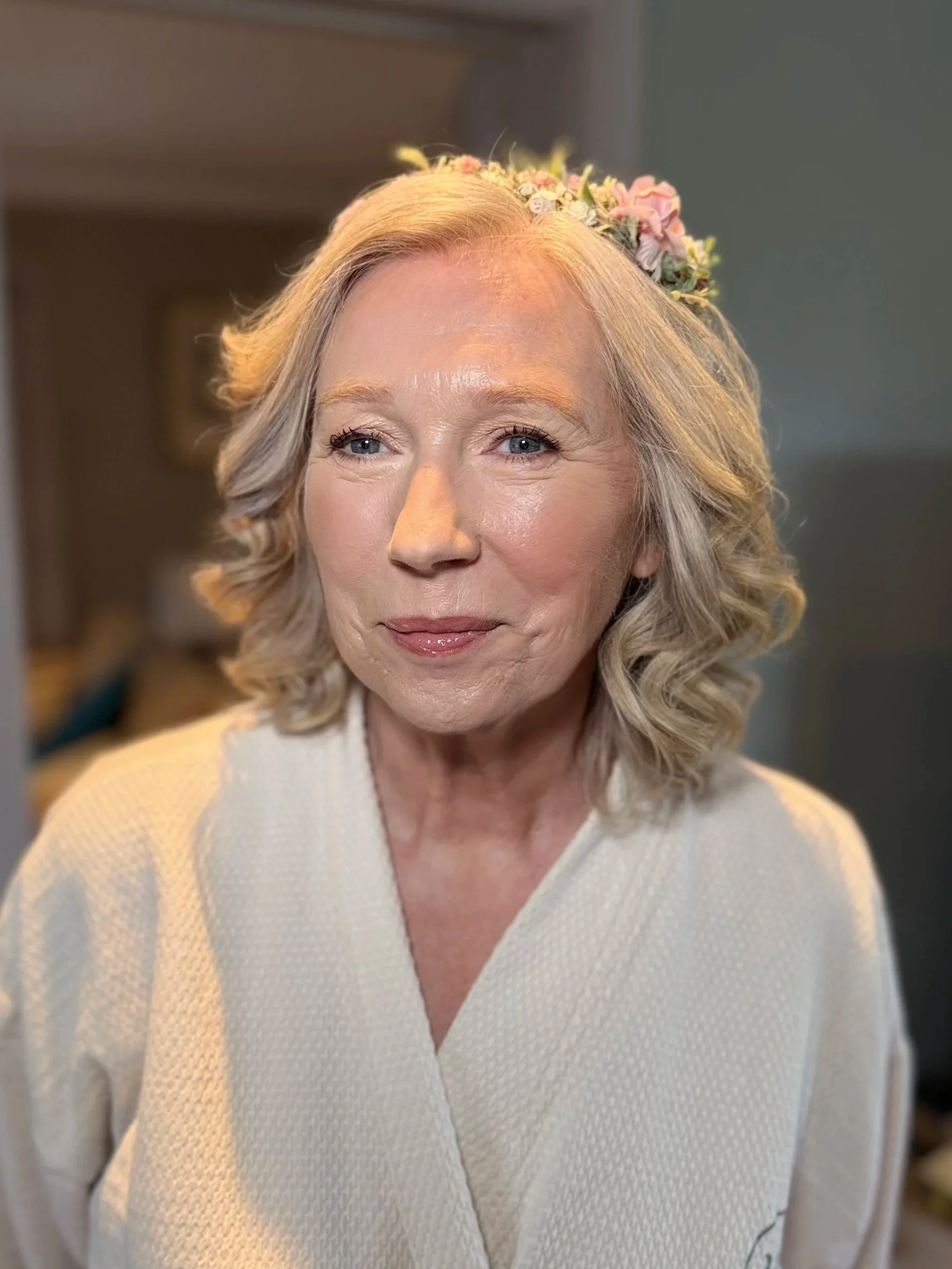 A smiling elderly woman with light blonde hair styled in soft curls, wearing a floral headband and a light-colored textured blouse, standing indoors.