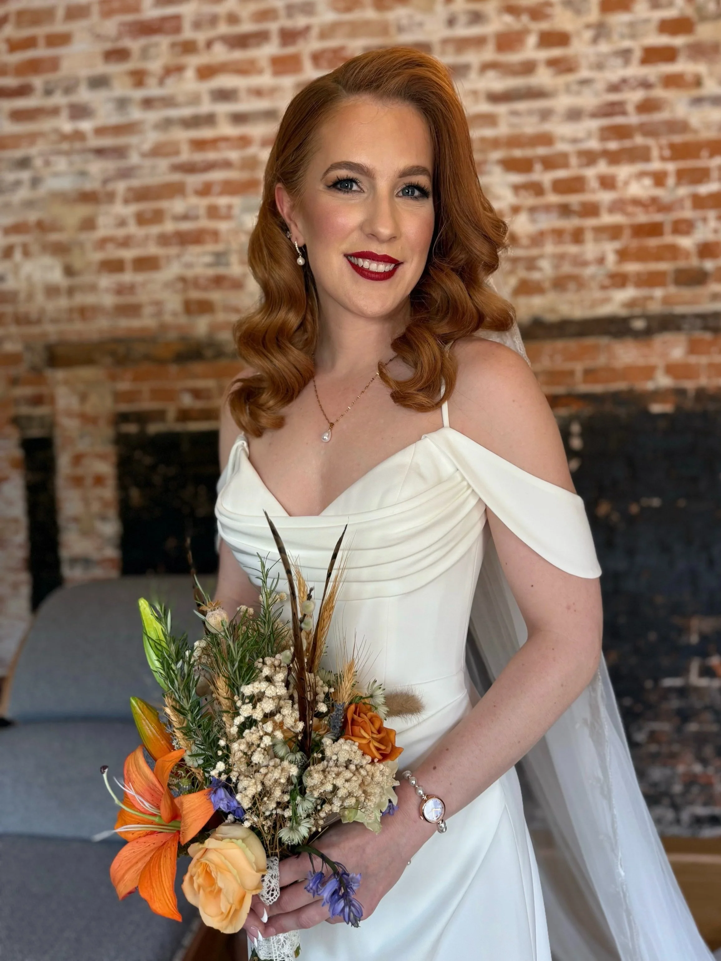 Bride with curly red hair in an off-shoulder white wedding dress, holding a bouquet with orange flowers against a brick wall background.