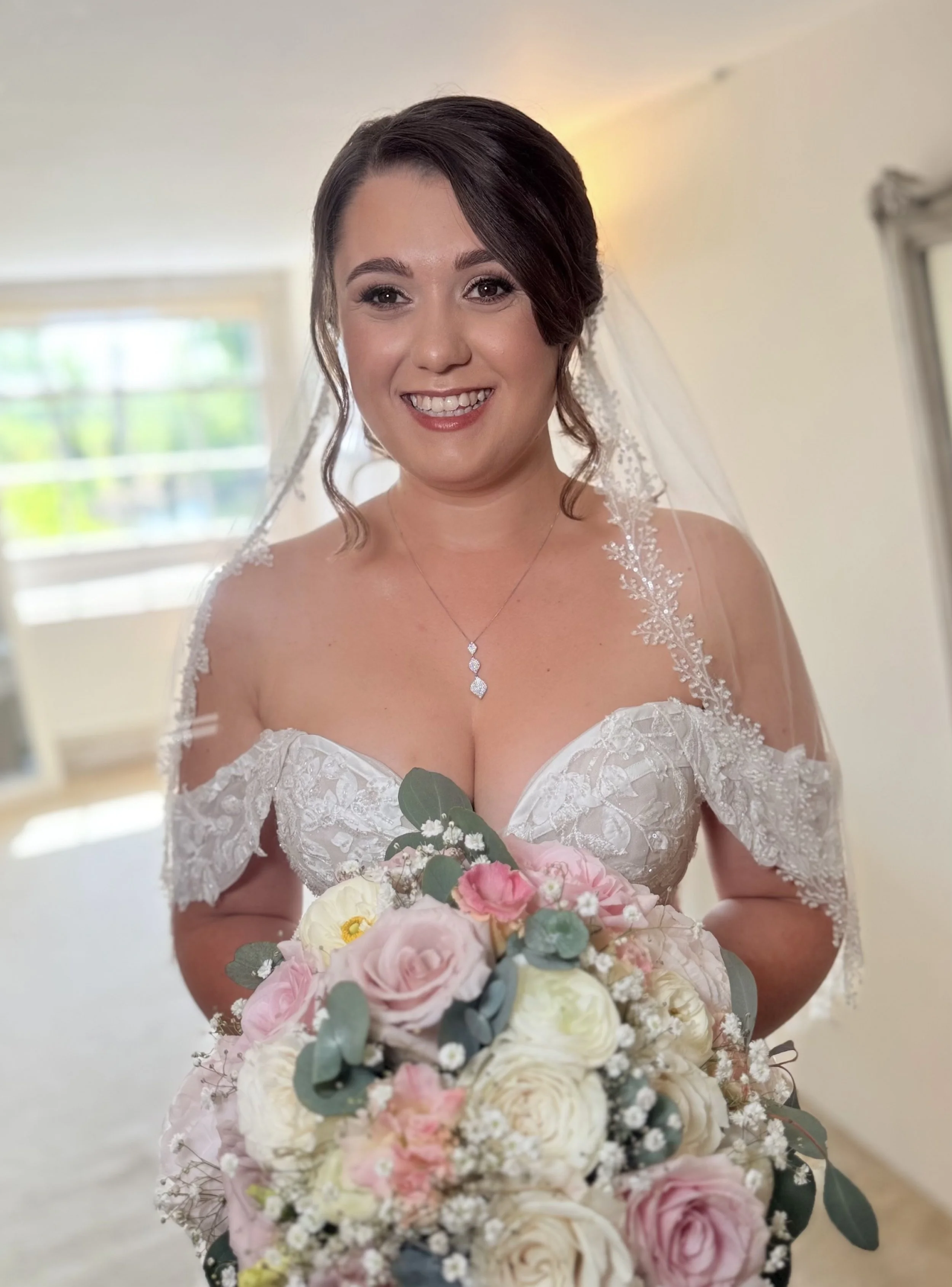 A bride in a white lace wedding dress holding a large bouquet of pink, white, and cream roses with greenery, smiling indoors with a window in the background.