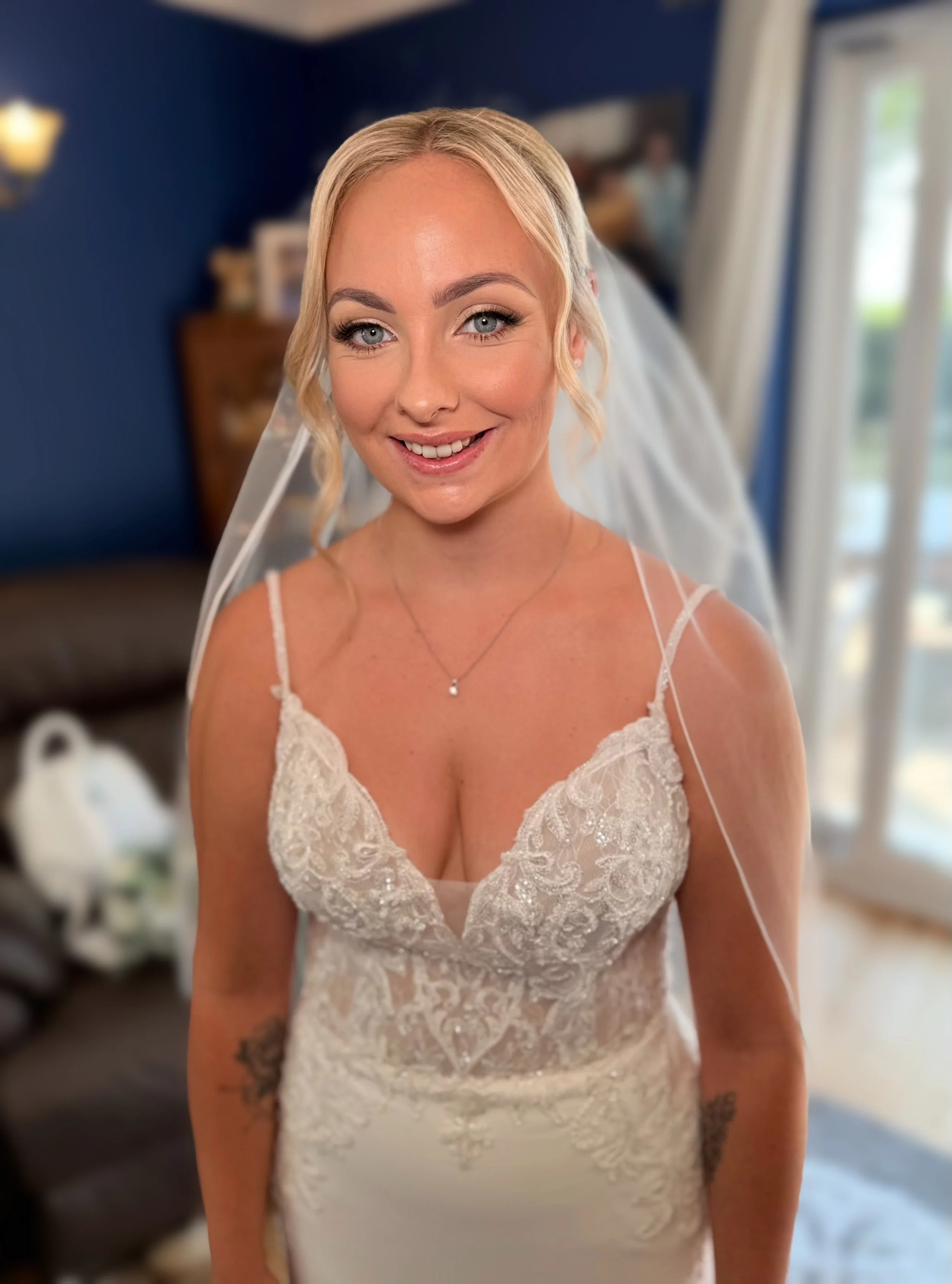 A bride in a white lace wedding dress with spaghetti straps, smiling in a room with blue walls and a window with white curtains.