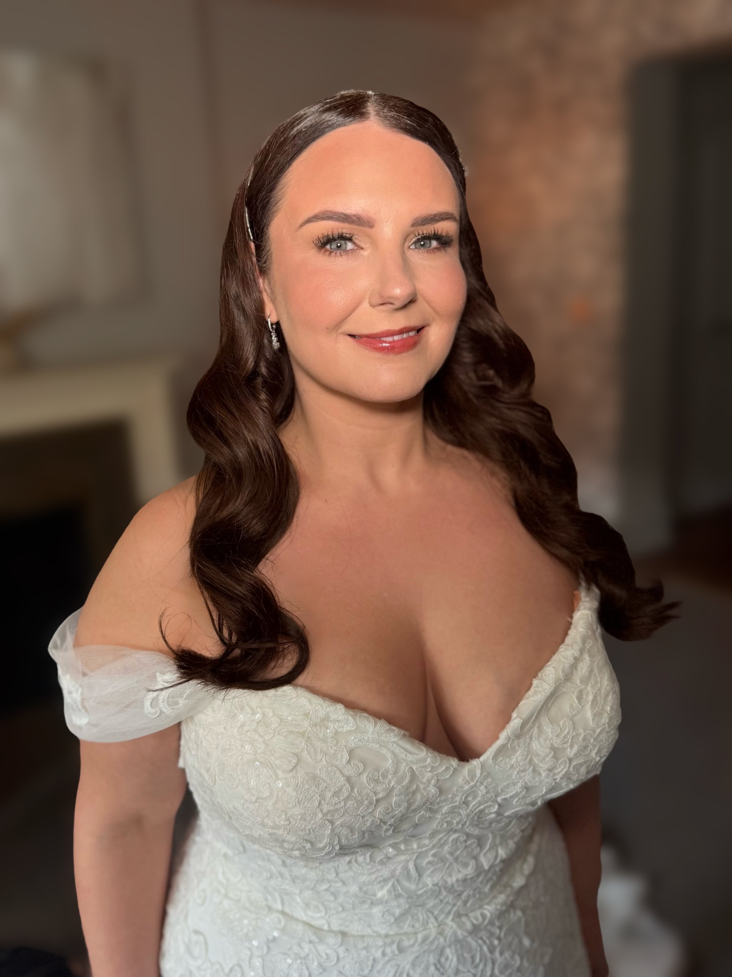 A woman with long wavy brown hair, blue eyes, and fair skin wearing a white lace off-the-shoulder dress, smiling in a softly lit indoor setting.