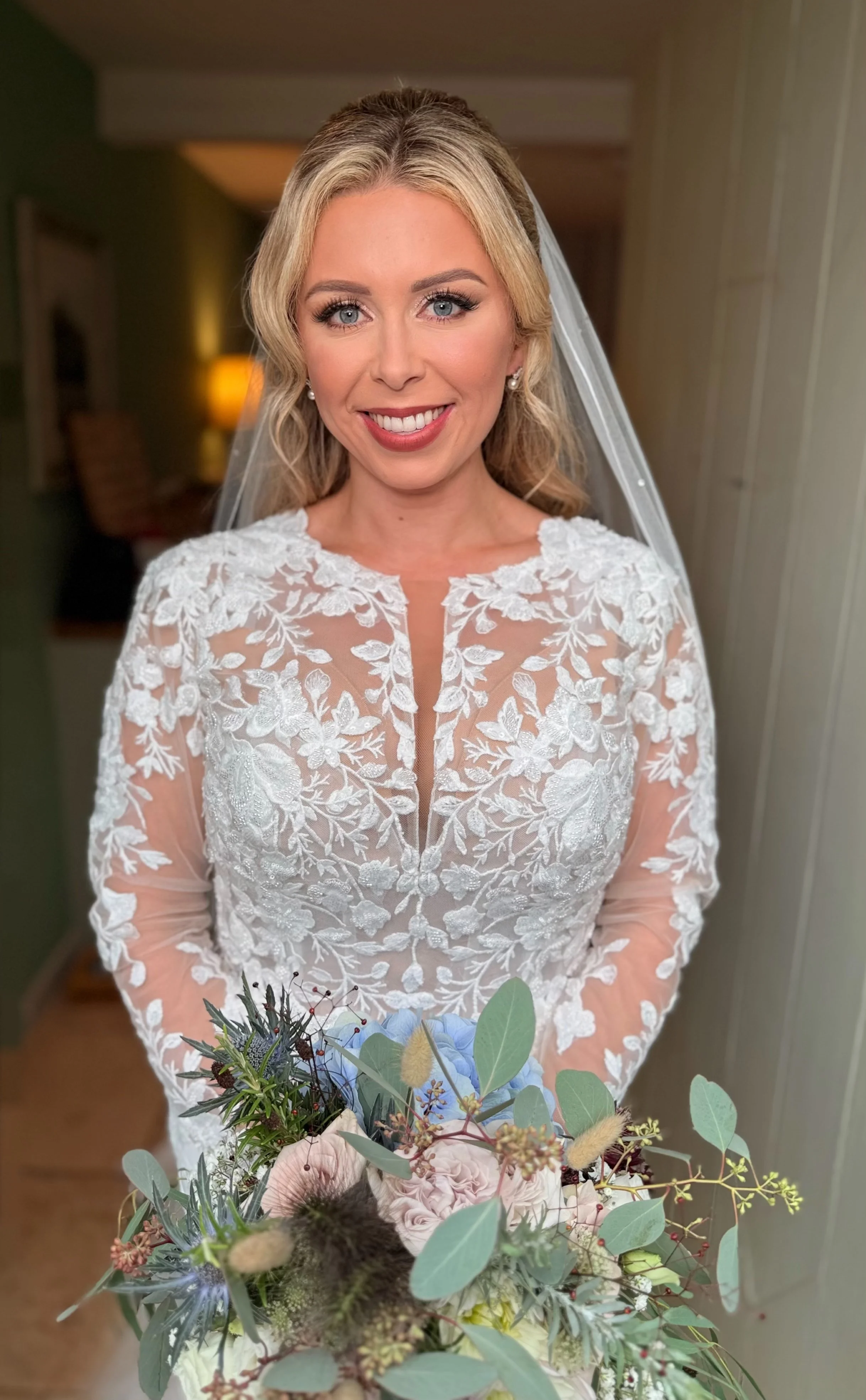 A smiling bride with blonde hair, wearing a white lace wedding dress and veil, holding a bouquet of flowers with roses, greenery, and other floral elements, standing indoors.