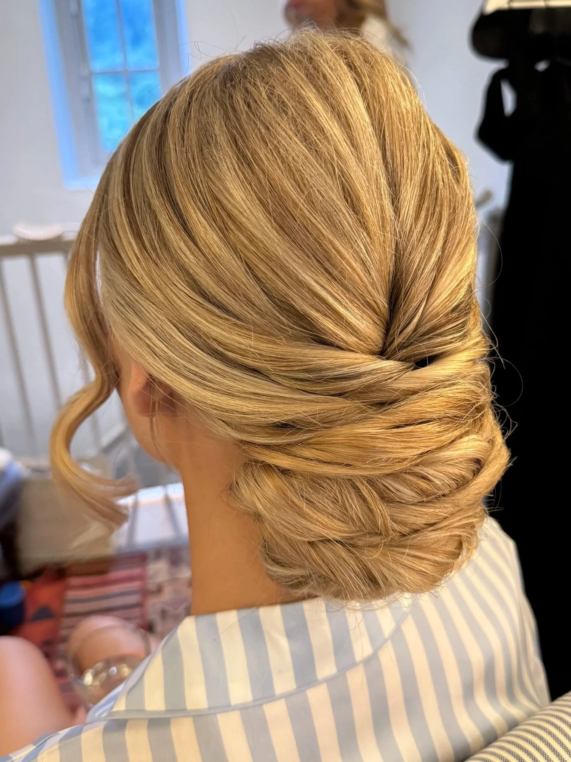 Back view of a woman with blonde hair styled in an elegant twisted updo. She is wearing a light blue and white striped shirt and is indoors near a window with natural light.