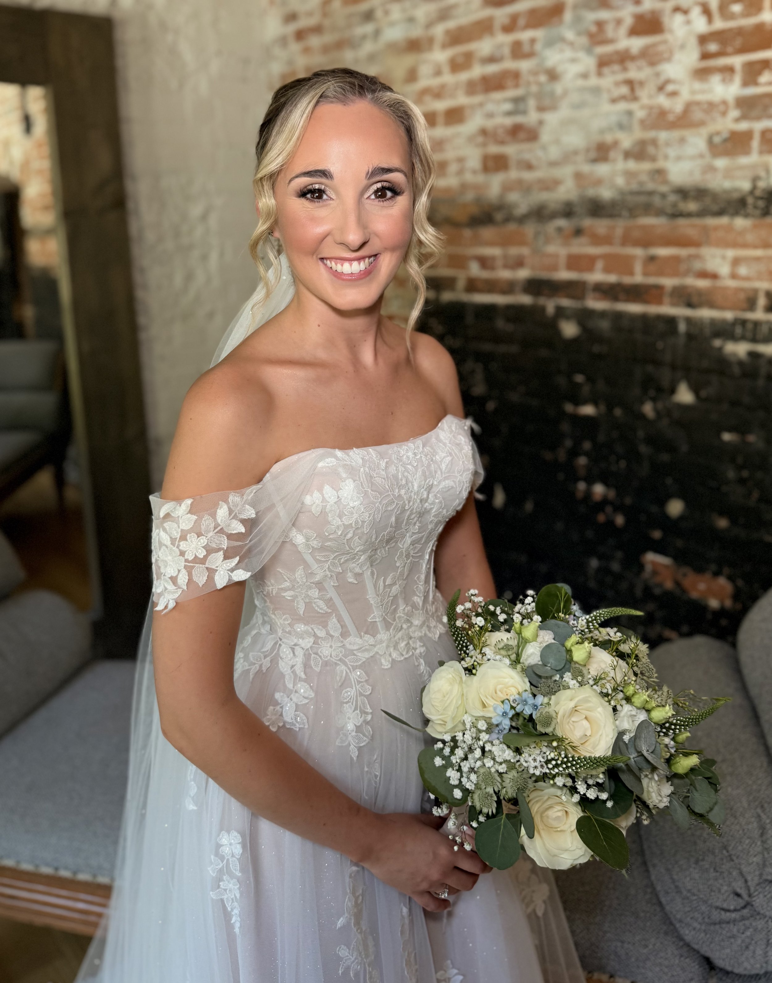A smiling bride wearing an off-the-shoulder white wedding dress holding a bouquet of white roses, baby's breath, and greenery, standing in front of a rustic brick wall.