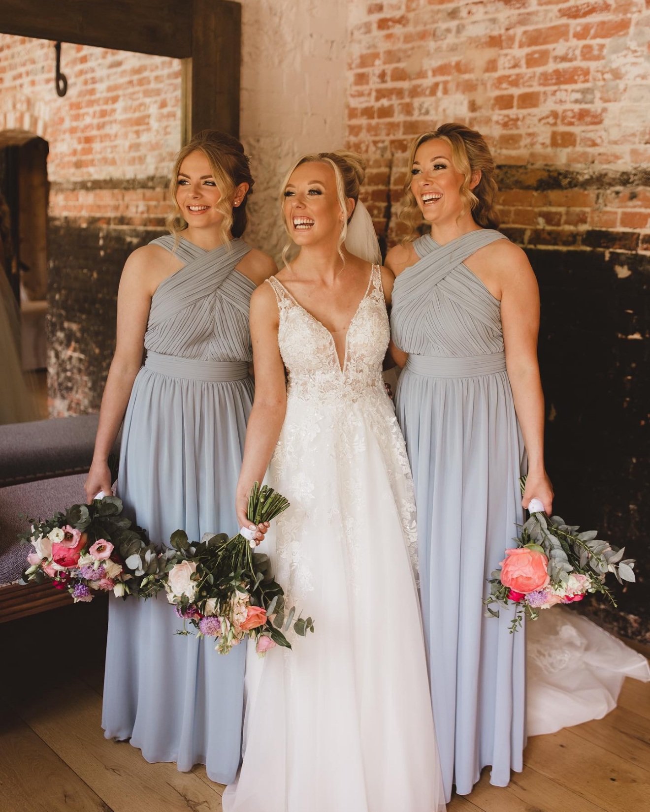 Bride and bridesmaids smiling, holding floral bouquets, in light blue dresses and white wedding gown, indoors with brick wall background.