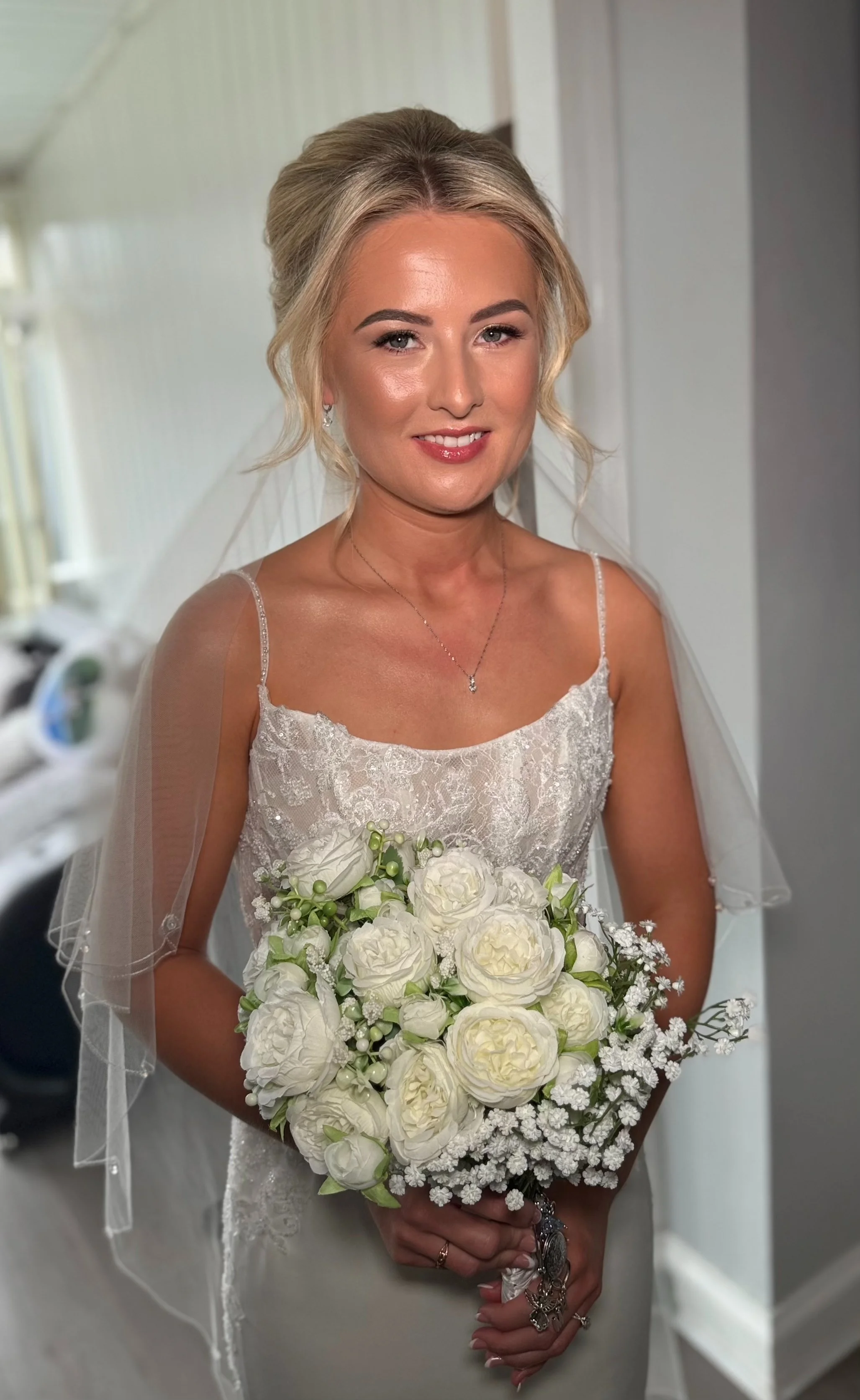 A bride with blonde hair in an updo, smiling, wearing a white lace wedding dress with spaghetti straps, holding a bouquet of white roses and greenery, standing indoors.