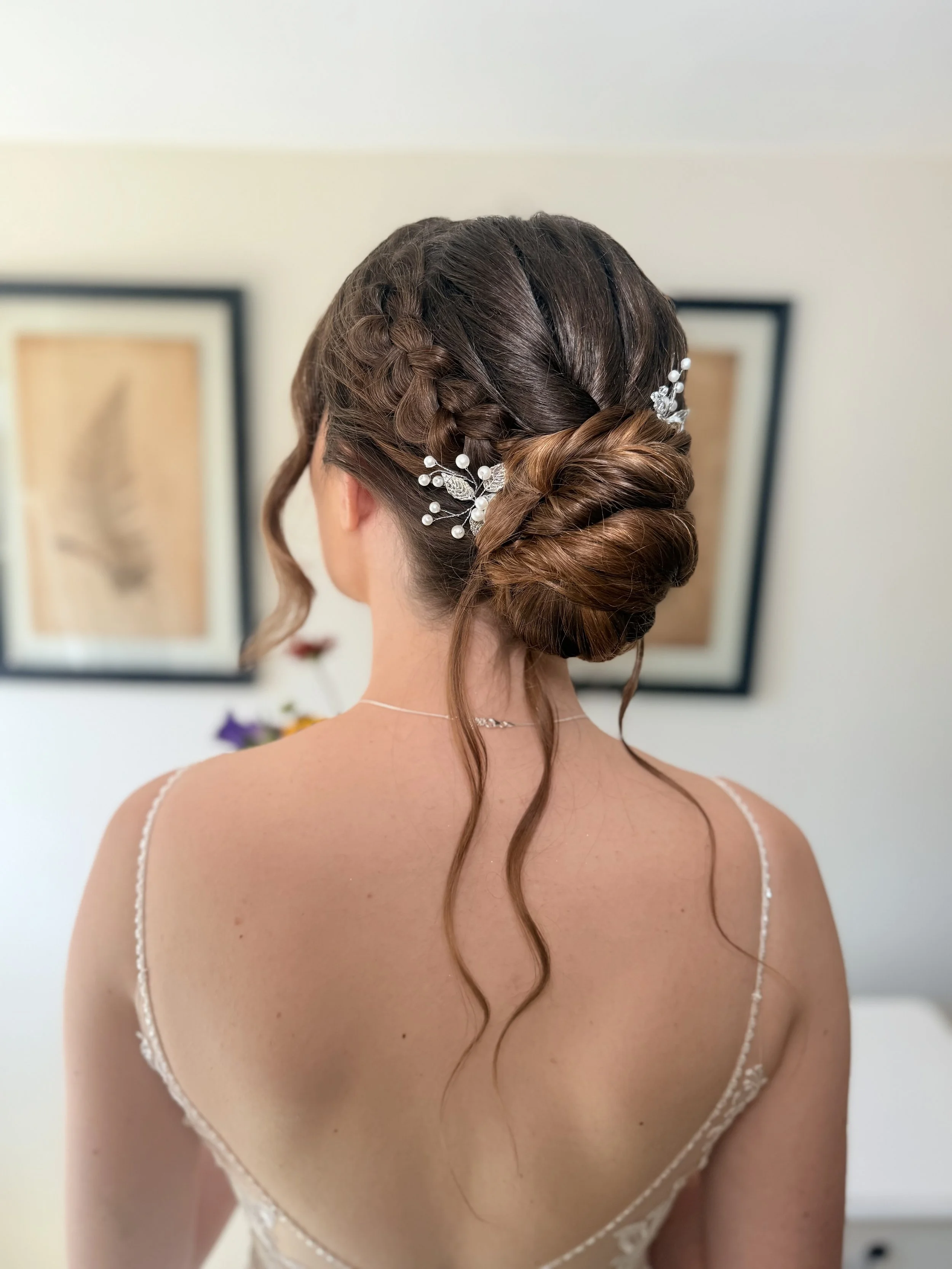 A woman with an elaborate braided updo hairstyle decorated with white pearl and crystal hair accessories, wearing a delicate spaghetti strap dress, standing indoors with framed artwork on the wall in the background.
