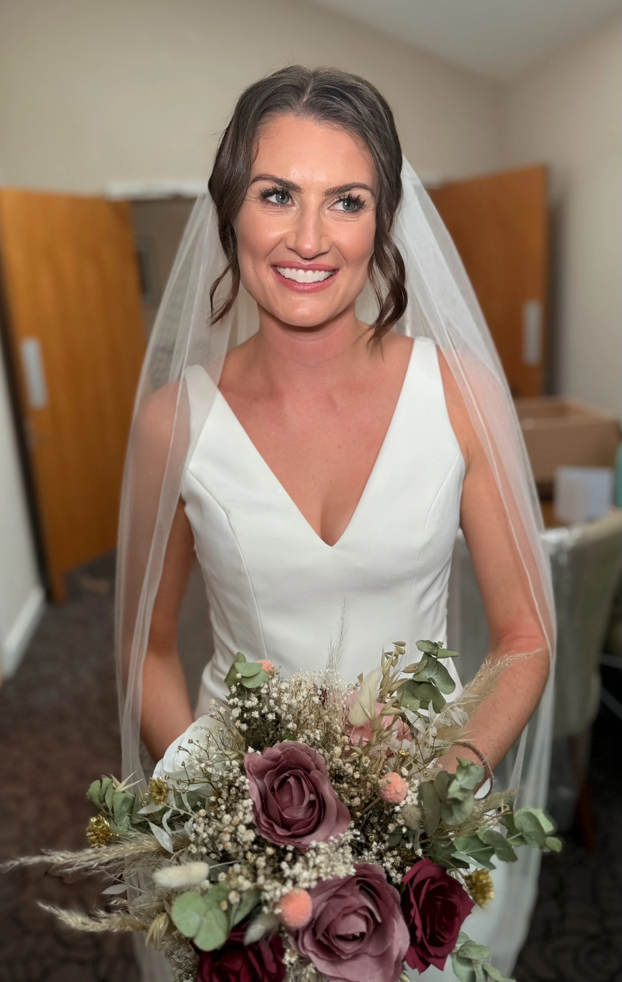 A bride in a white dress holding a bouquet of roses and other flowers, smiling, and wearing a veil.