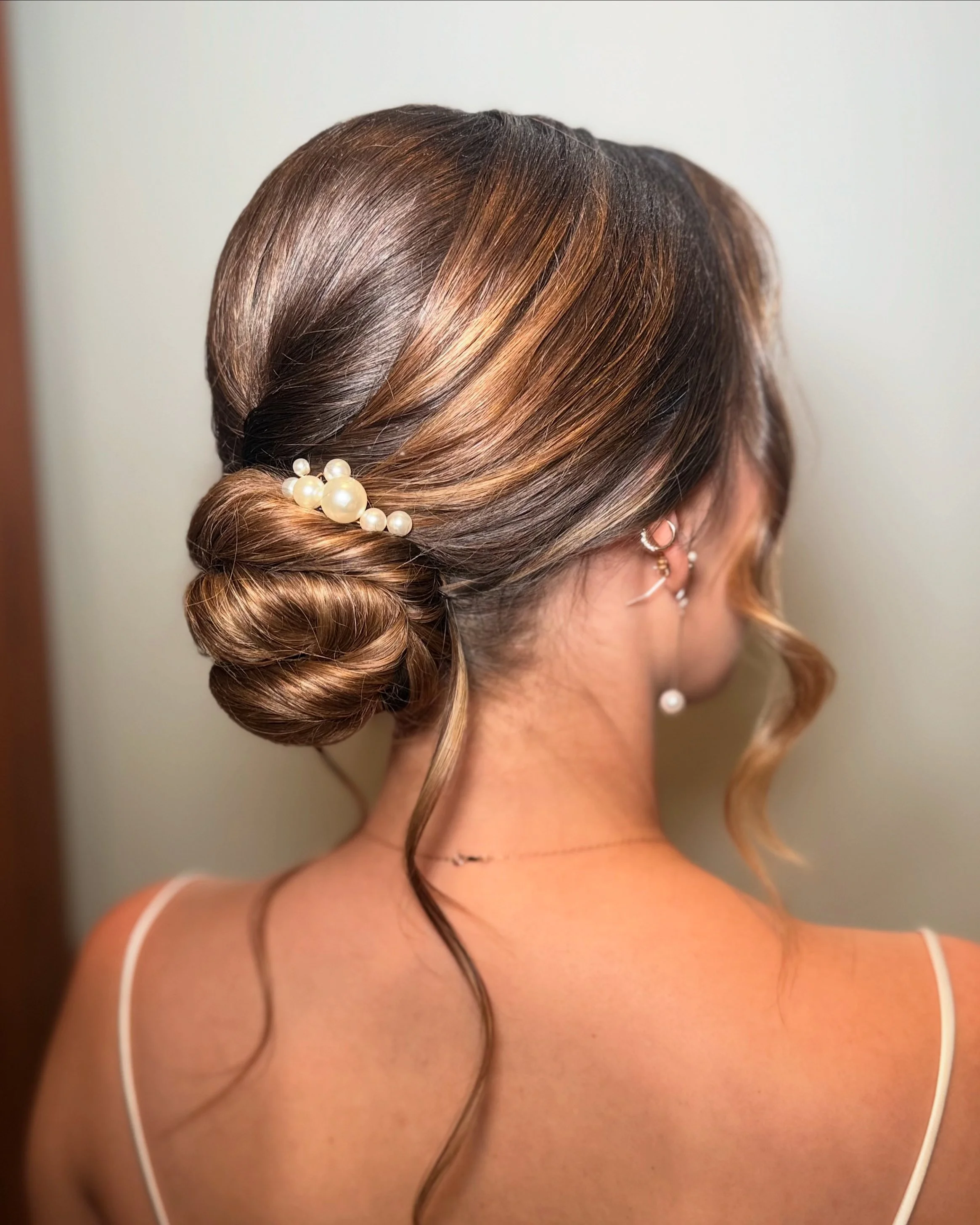 Back view of a woman's hair styled in an elegant bun with a pearl hair accessory, wearing pearl earrings, and a thin-strapped top.