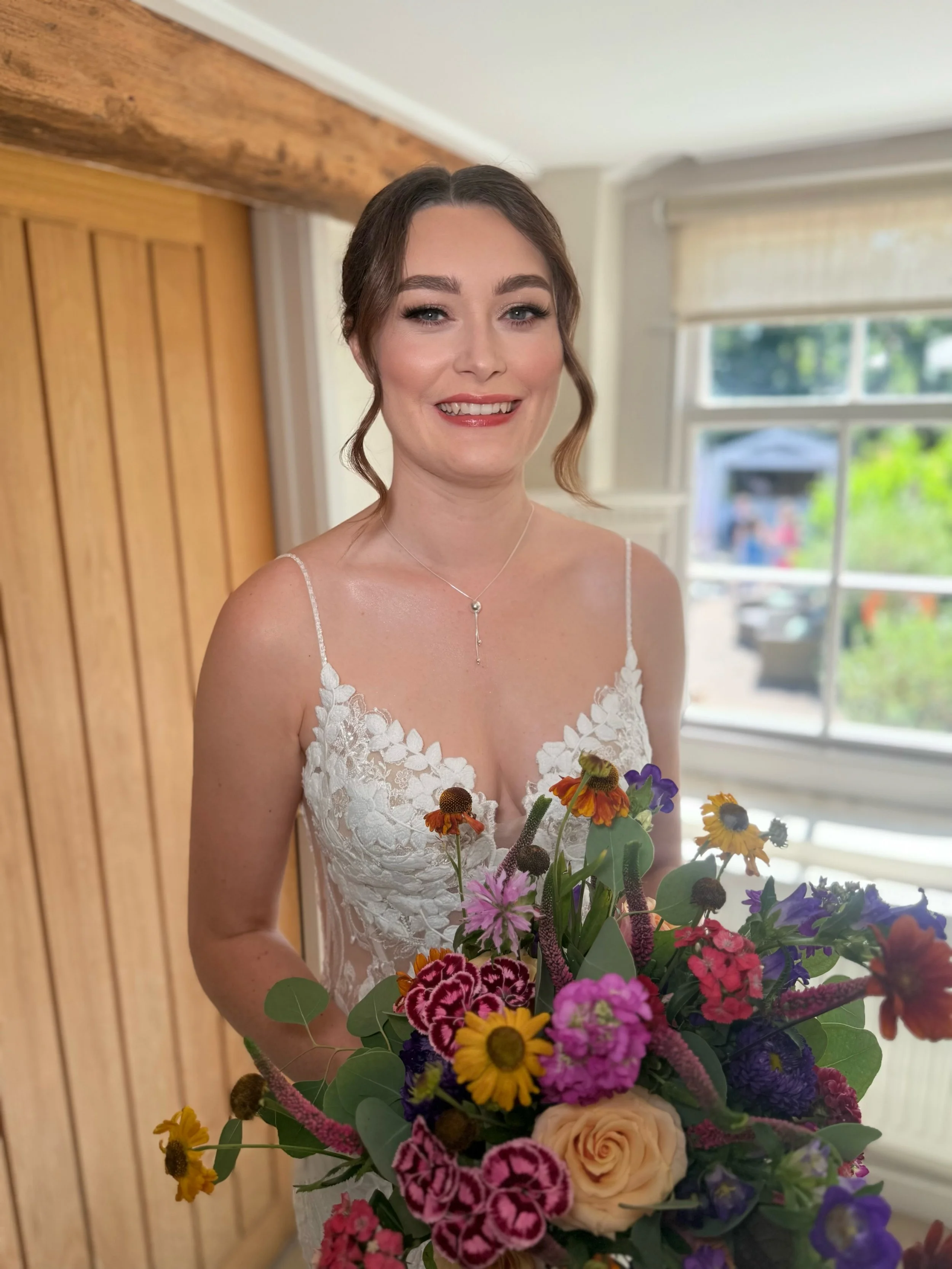 A smiling woman in a bridal gown with lace detail, holding a colorful bouquet of flowers indoors near a window.