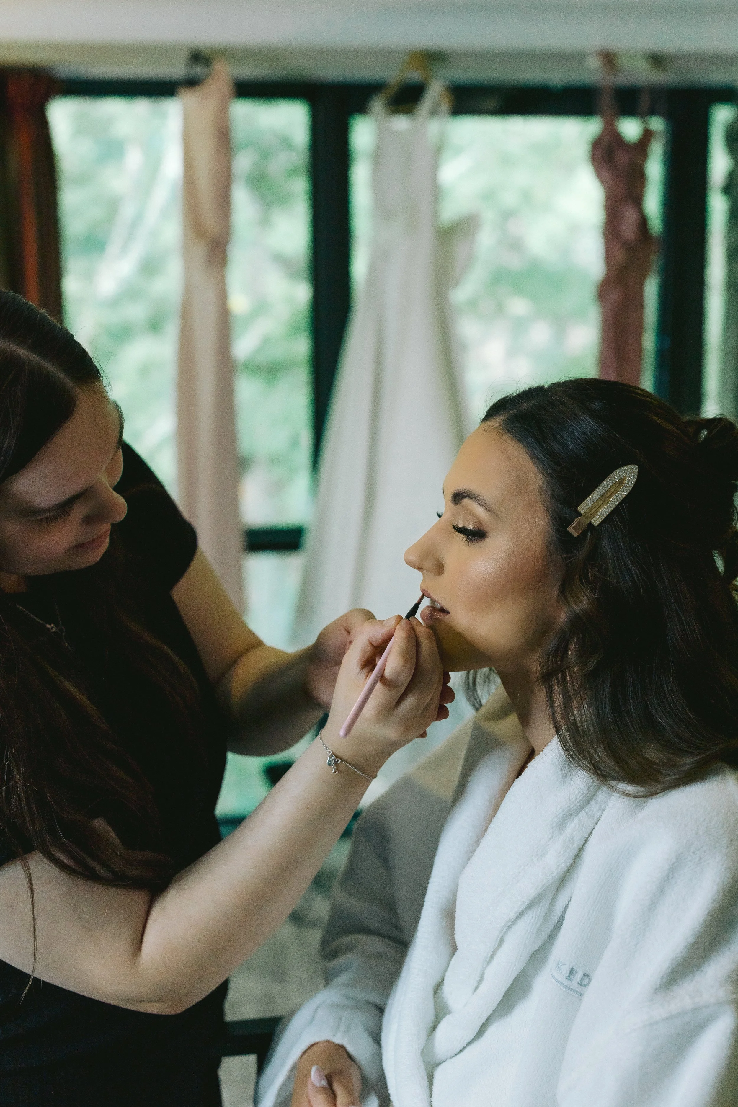 Makeup artist applying lipstick to a woman in a white robe, with dresses hanging in the background.