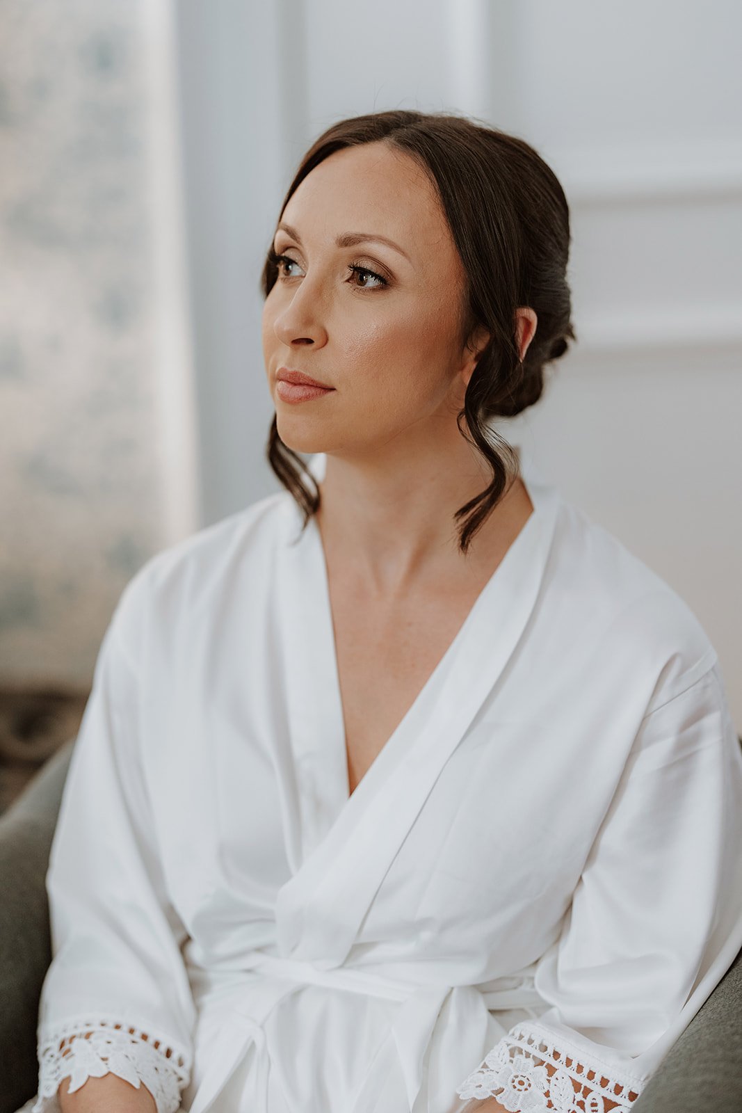 A woman in a white robe, with her hair styled in an updo, looking thoughtfully to the side in a softly lit room.