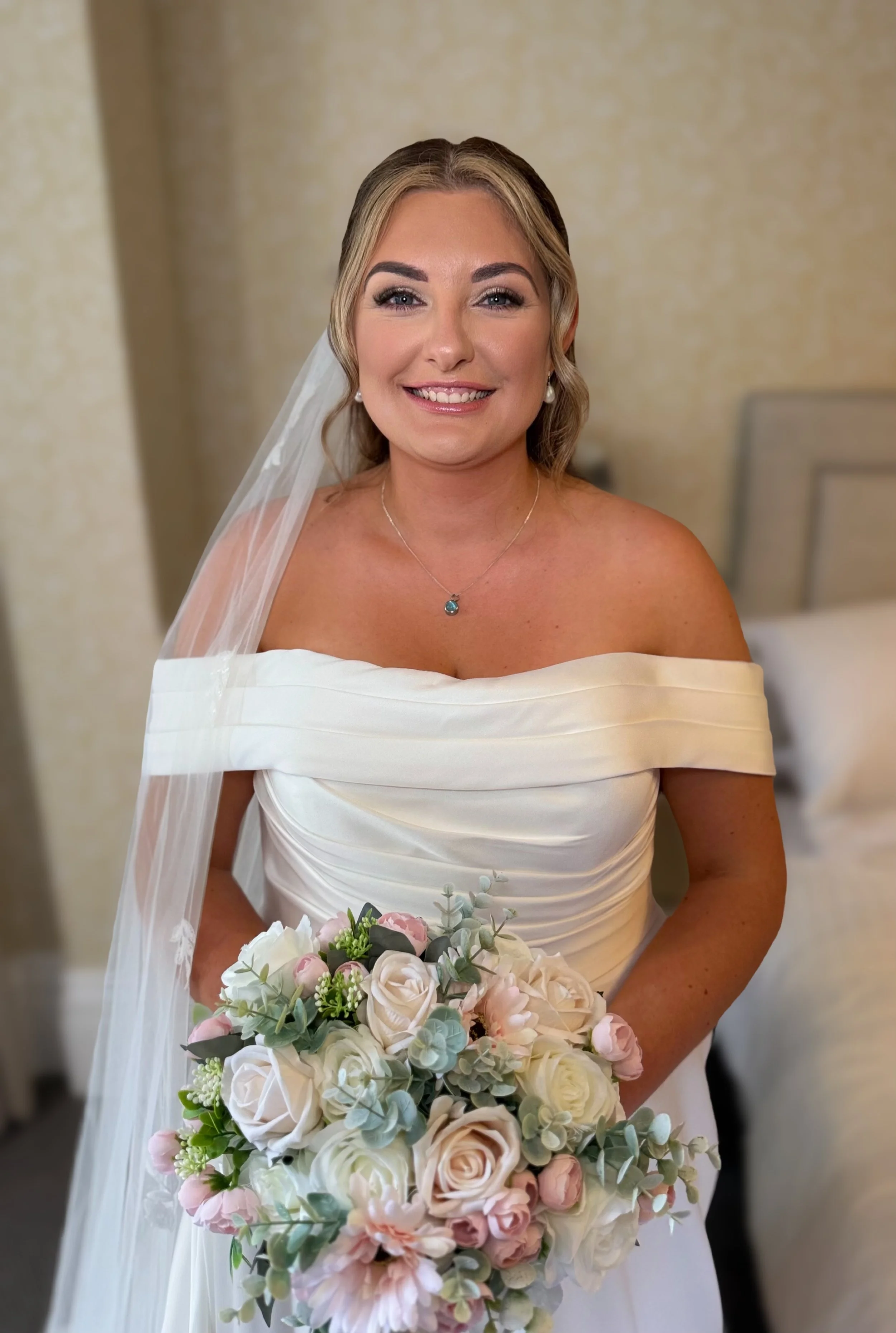 Smiling woman in a wedding dress holding a bouquet of white and pink roses, wearing pearl earrings and a necklace, with a veil on her hair, in a softly lit room.