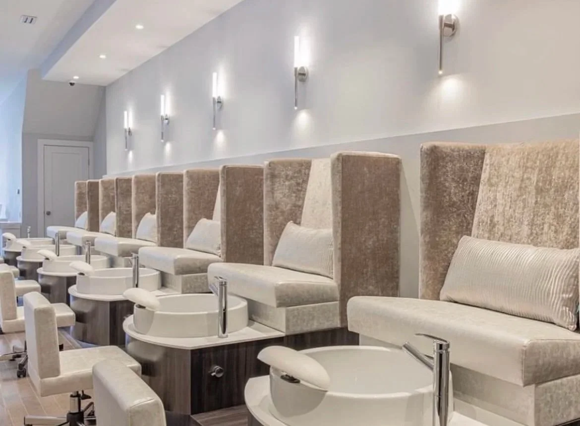 A row of beige upholstered pedicure chairs with foot baths at a salon, with wall-mounted lights above.