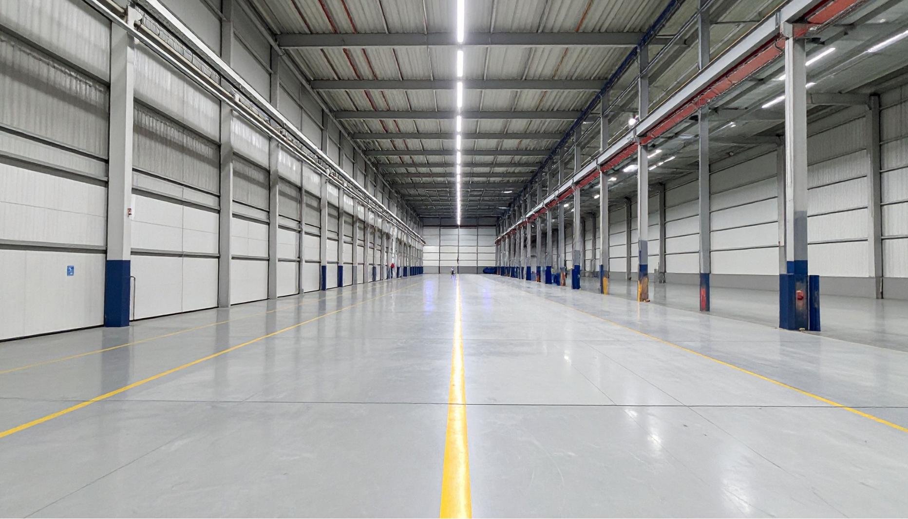 Empty warehouse with high ceilings, gray concrete floor, bright overhead lighting, white walls, and large grid-style doors.