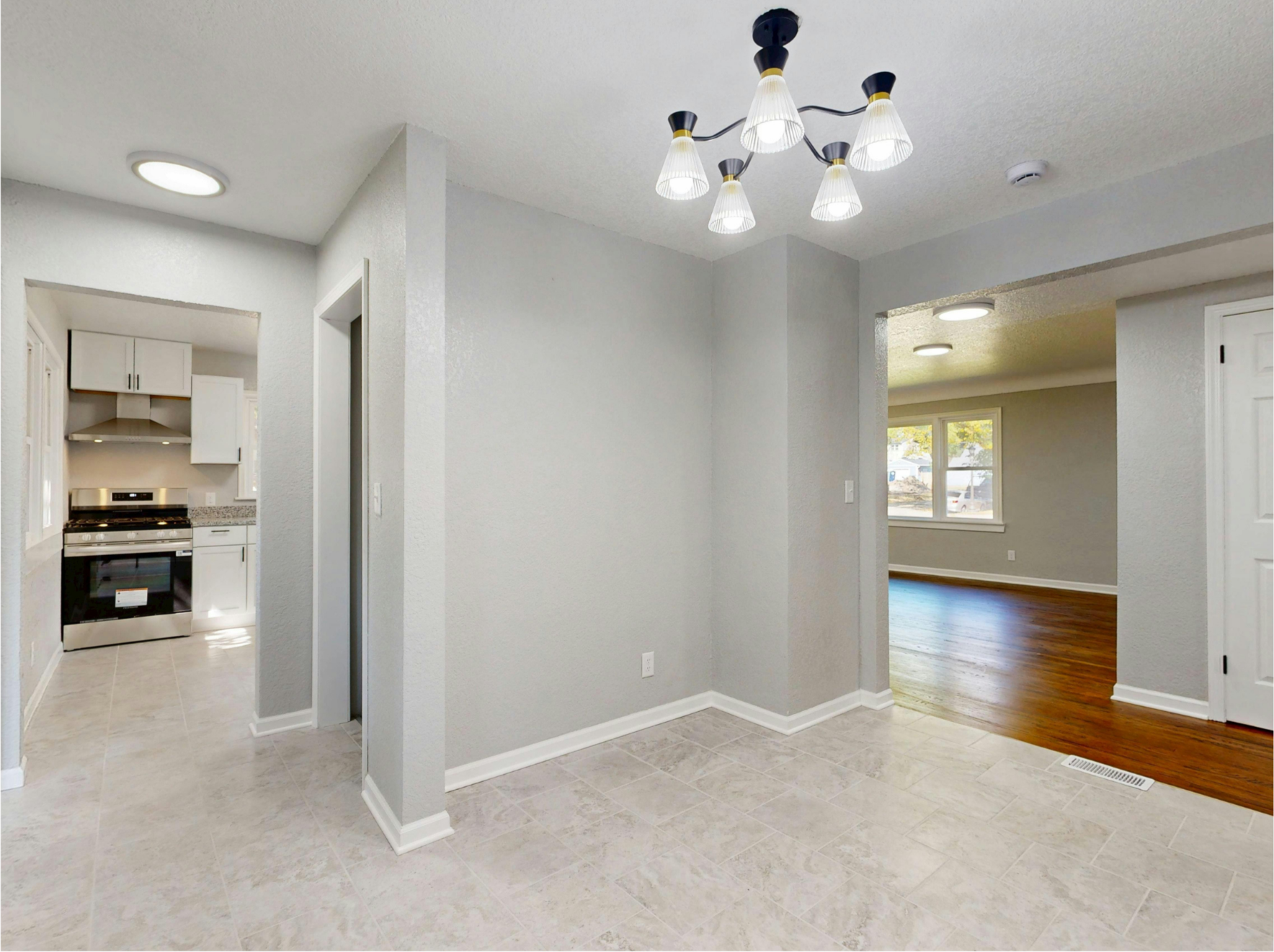 Empty room with light-colored tiled flooring, gray walls, a ceiling chandelier, and an adjoining room with dark wood flooring and a big window.