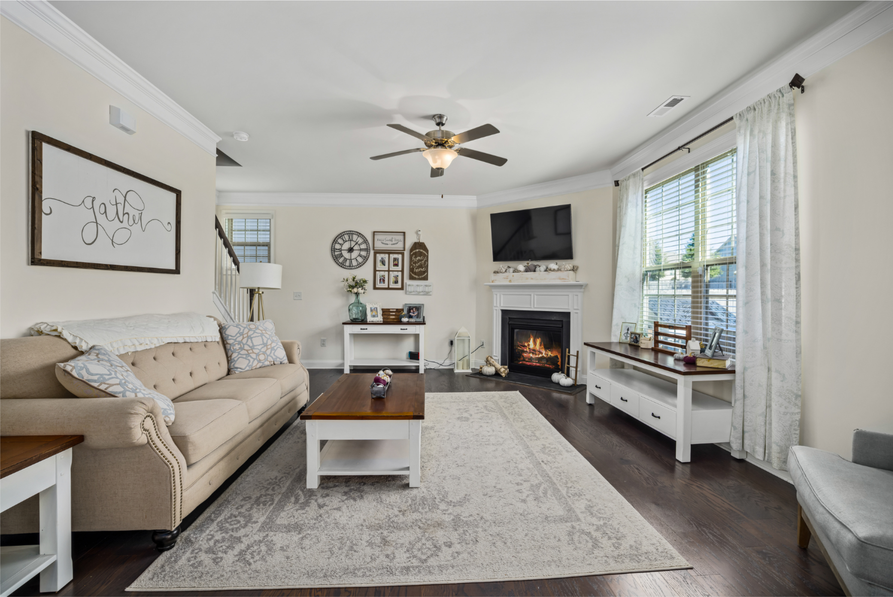 Living room with beige couch, fireplace, wall-mounted TV, dark hardwood floors, window with curtains, area rug, coffee table, side tables, wall decor, and decorative accents.