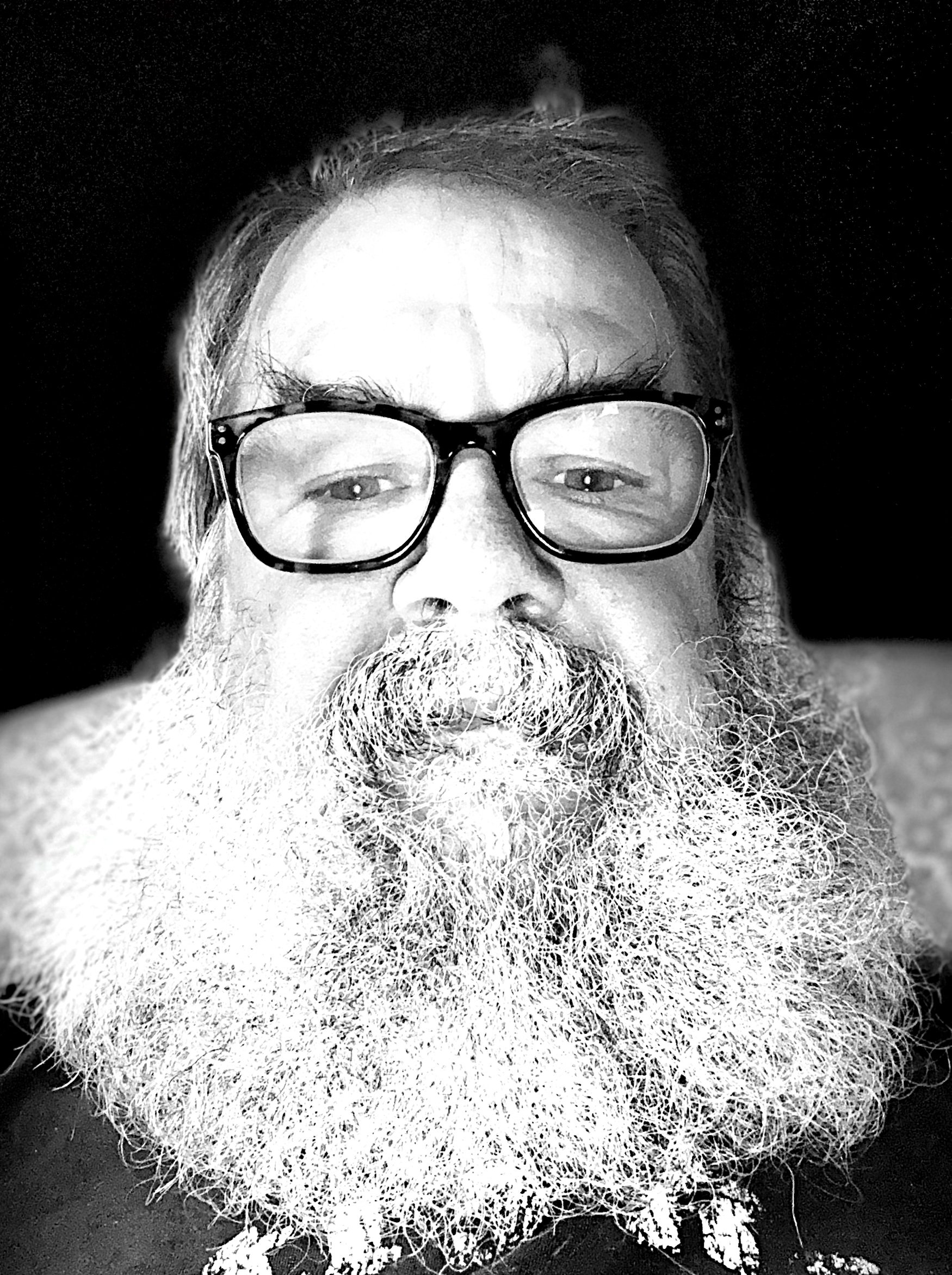 Black and white close-up of a man with glasses, a full white beard, and long hair, sitting against a dark background.