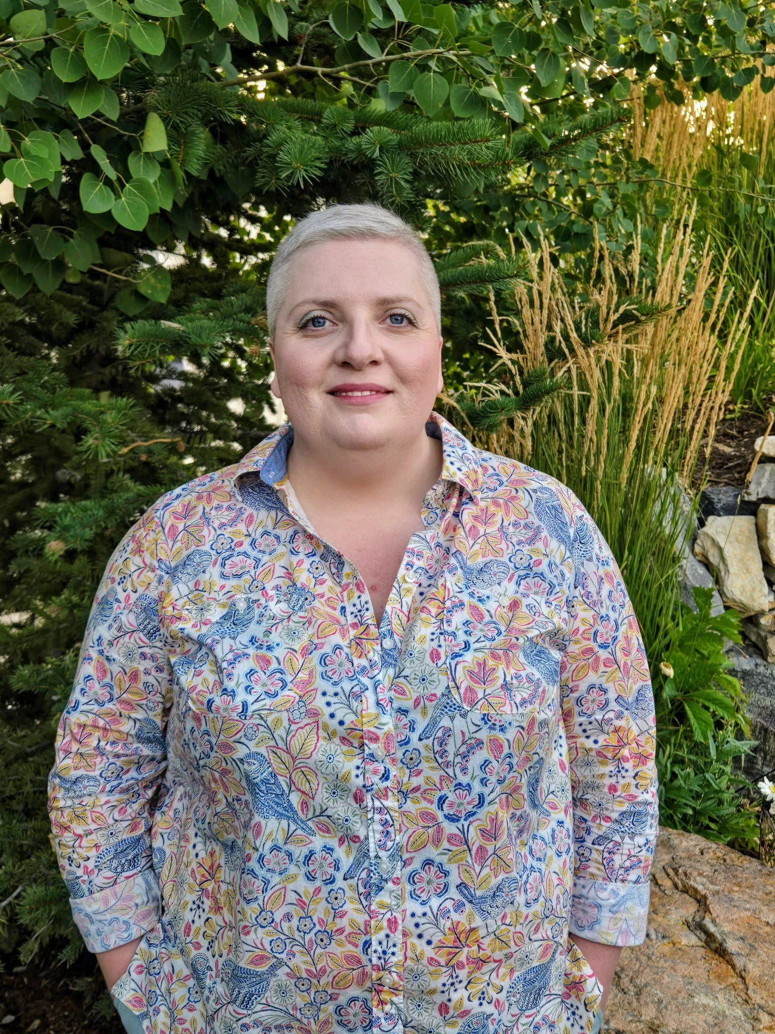 A person with short blonde hair and blue eyes standing outdoors in front of green foliage and plants, wearing a colorful floral patterned shirt.