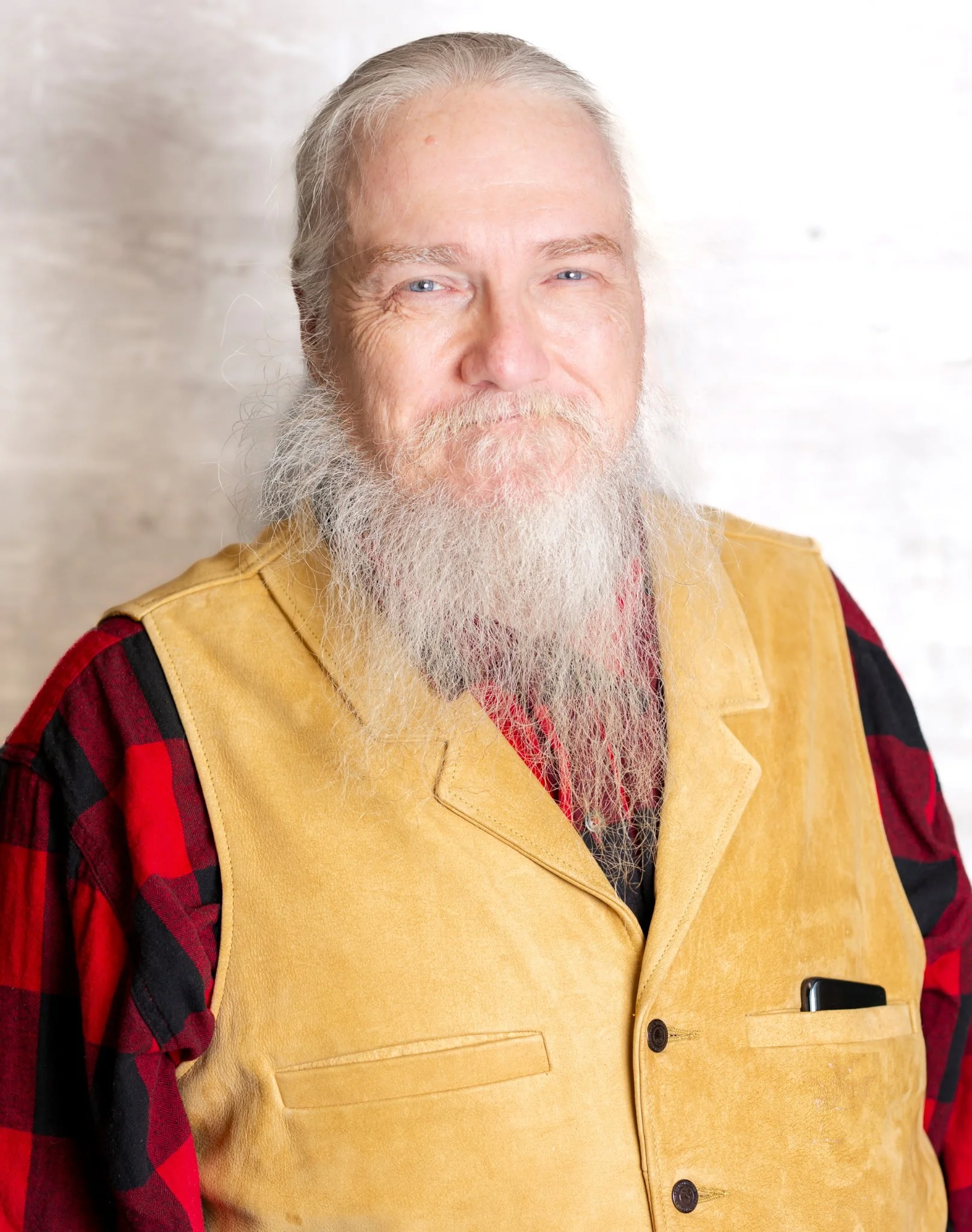 A portrait of an older man with a long white beard and white hair, wearing a yellow vest over a red and black checkered shirt, smiling gently.
