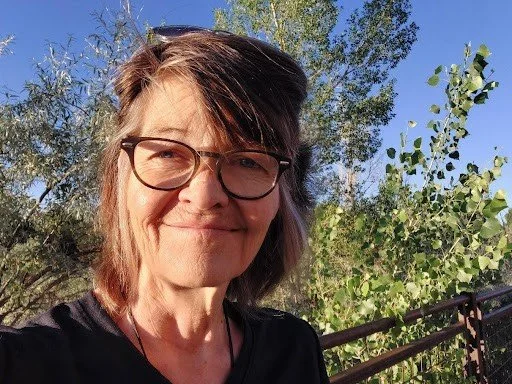 A woman with short brown hair, glasses, and wearing a black shirt smiling outdoors with trees and a clear blue sky in the background.