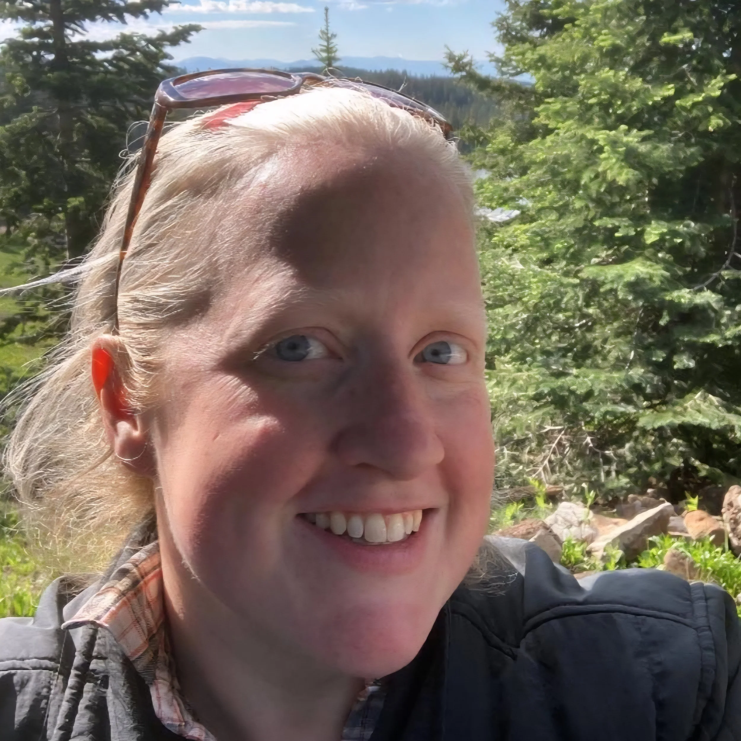 Close-up of a smiling woman with light hair and blue eyes outdoors, wearing sunglasses on her head, greenery, trees, and mountains in the background.