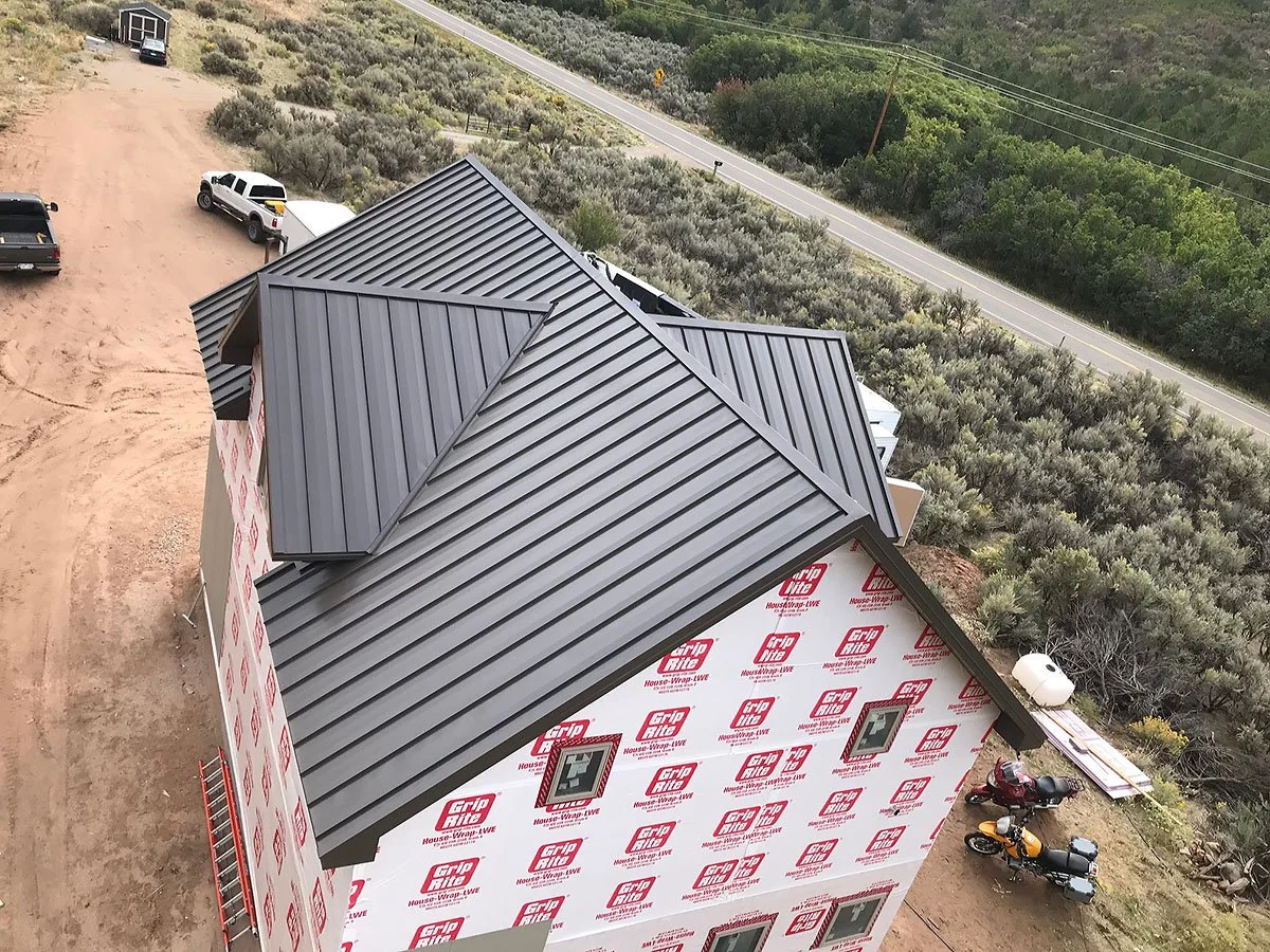 A house under construction with new black metal roofing, pink and white house wrap, three small windows, parked motorcycles, and surrounded by a dirt yard and natural greenery near a rural road.