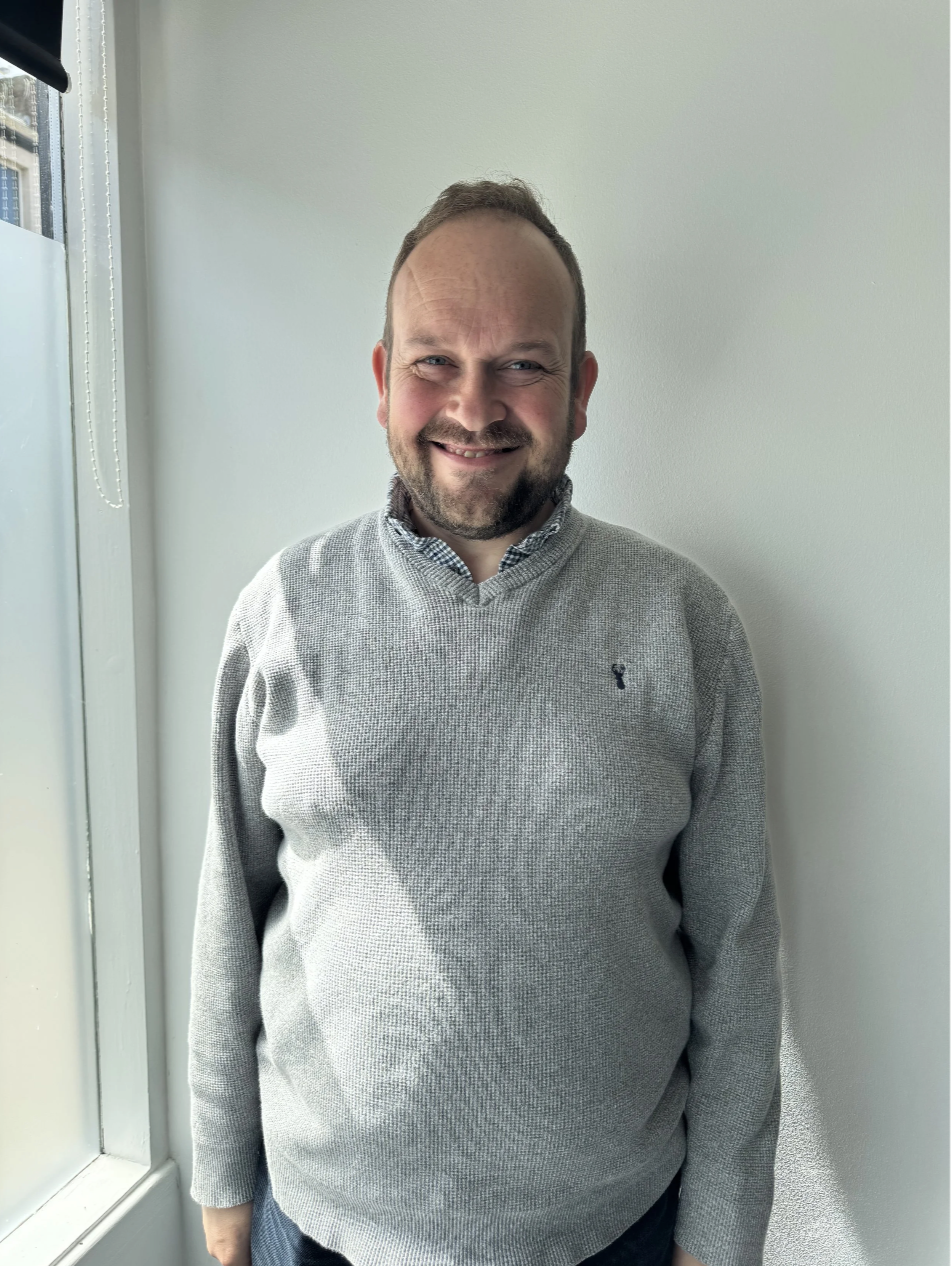 A smiling person, with short brown hair, wearing a light grey jumper. Standing in front of a white wall.
