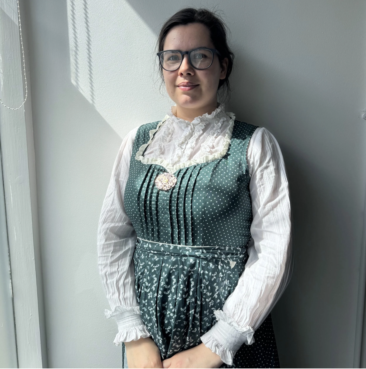 A person with brown hair that is tied back, wearing glasses, a dark green, floral dress, with a white long sleeved top underneath. They are standing in front of a white wall.